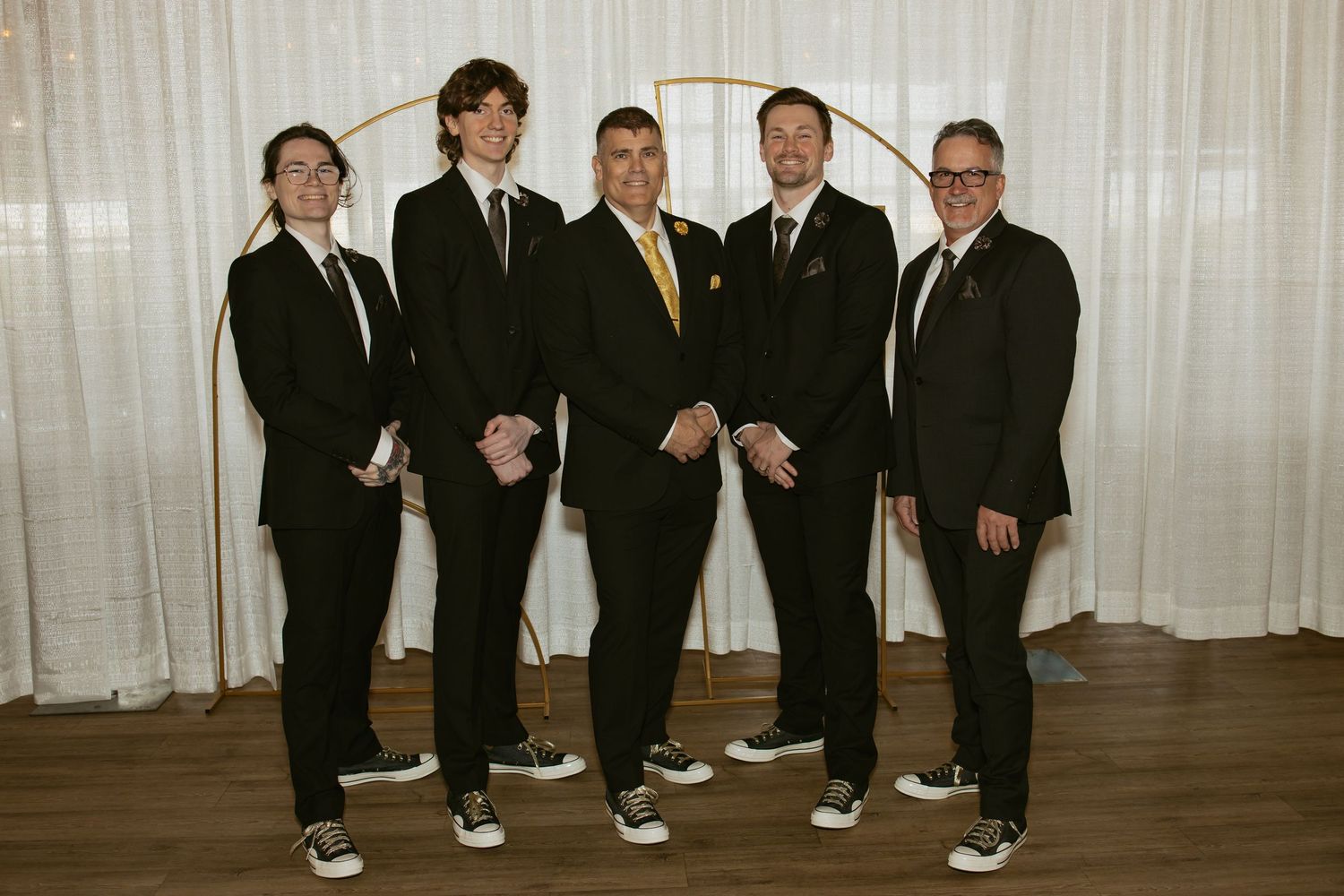 Five men in matching black suits pose together smiling at a formal event near a gold arch backdrop.