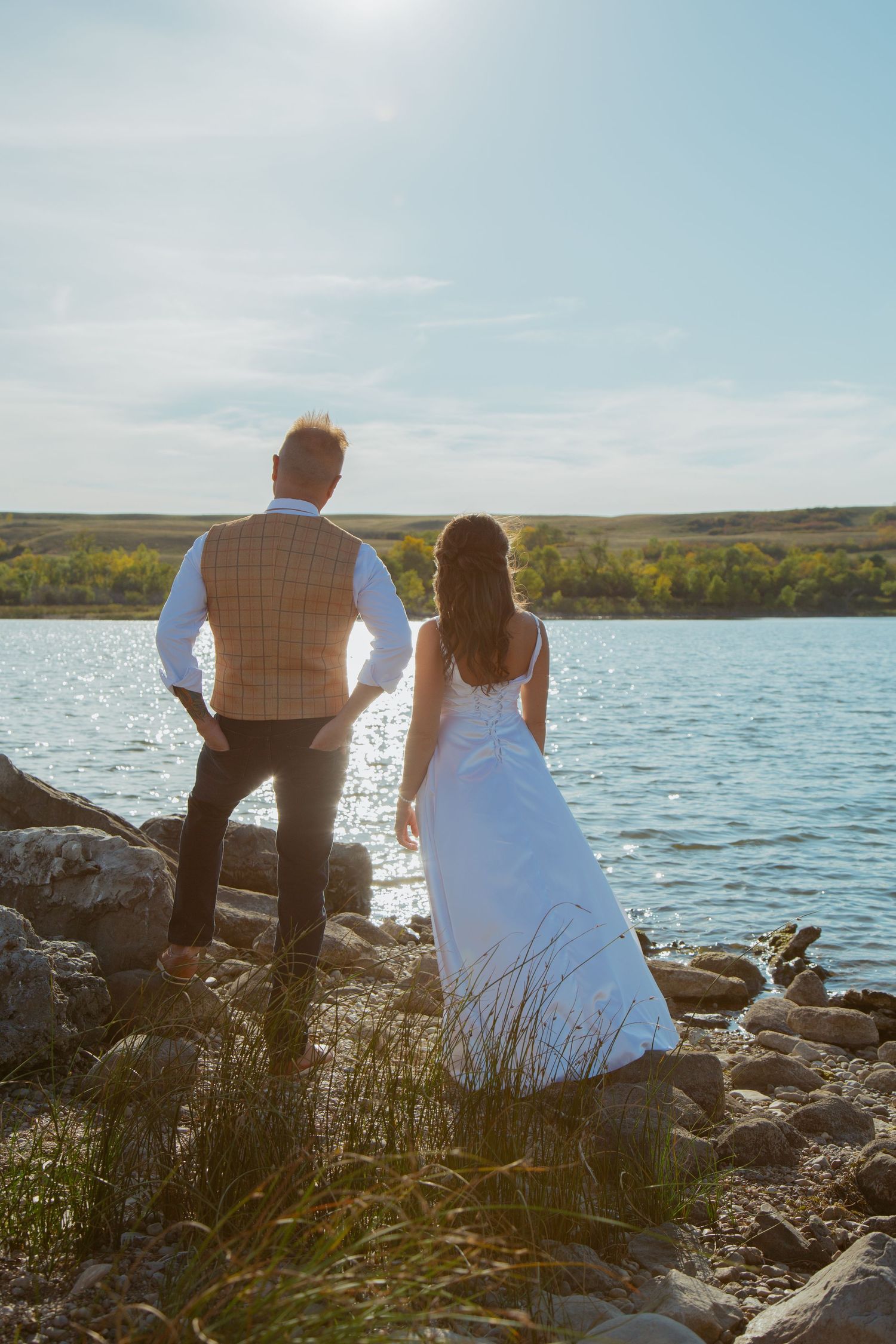 Couple stands on rocky shoreline overlooking serene lake at sunset.