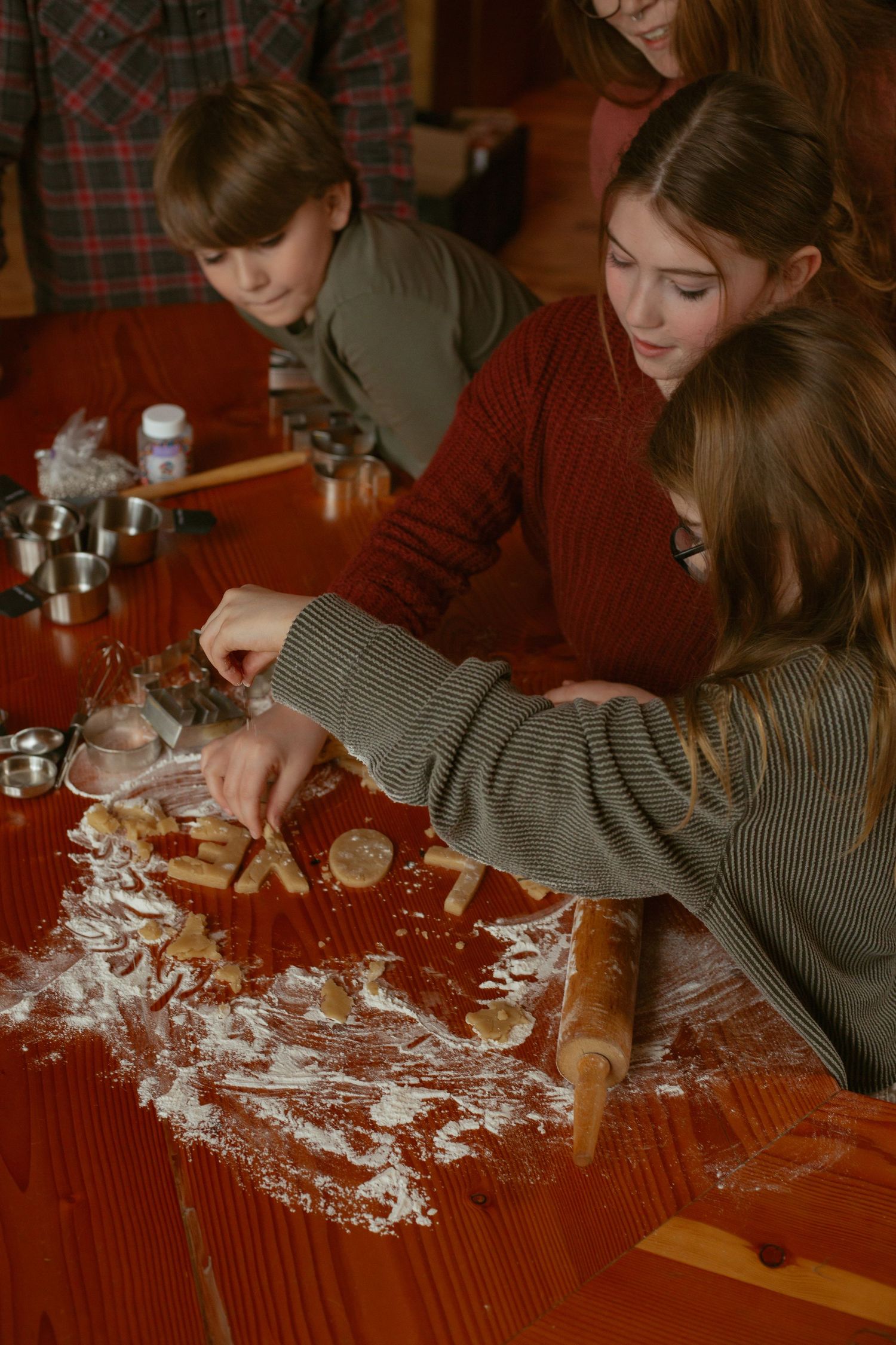 Children and adult cutting holiday cookies with flour-dusted red wooden table and rolling pin during festive baking.