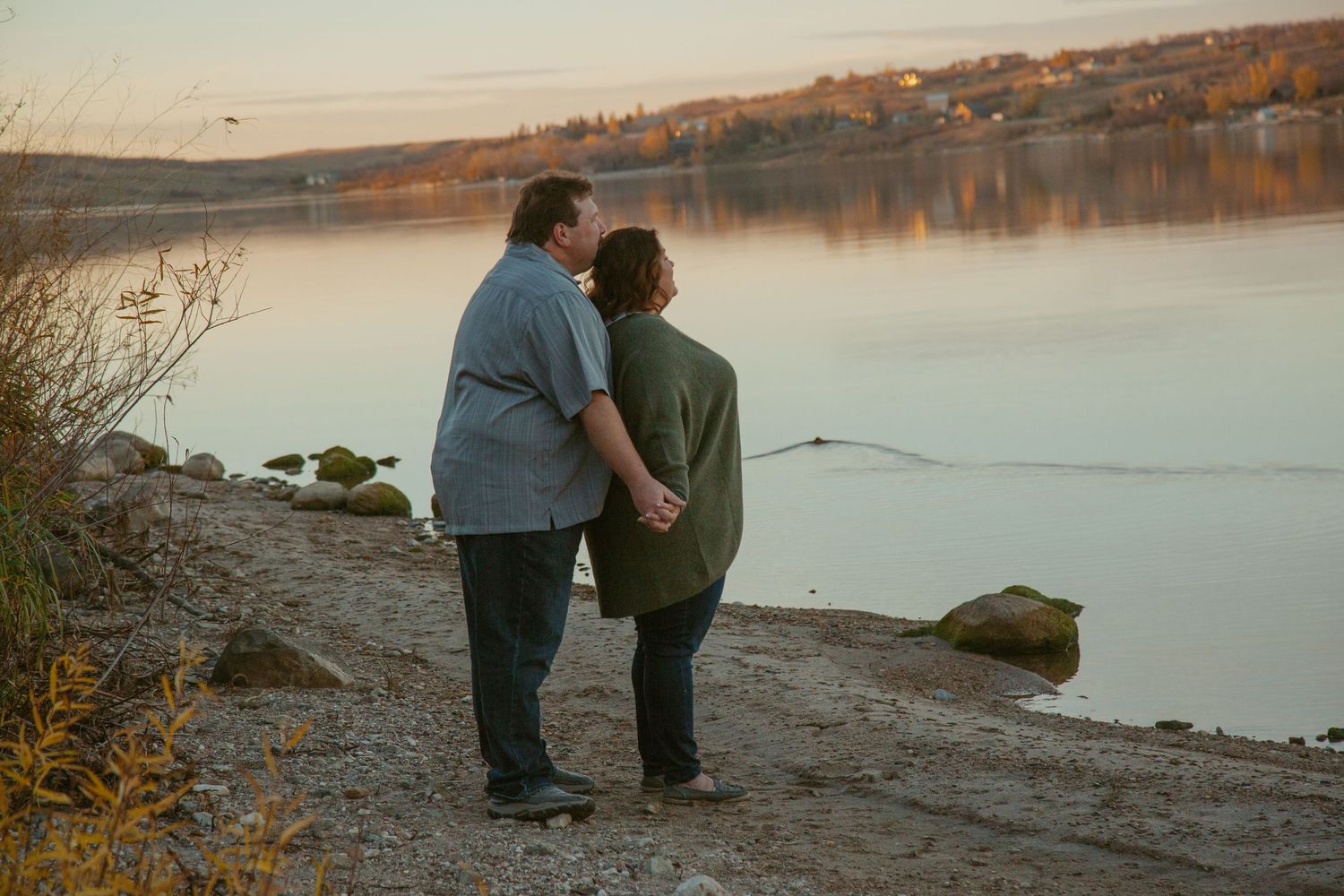 Couple walking along a scenic lakeside beach at sunset with mountains in the background.