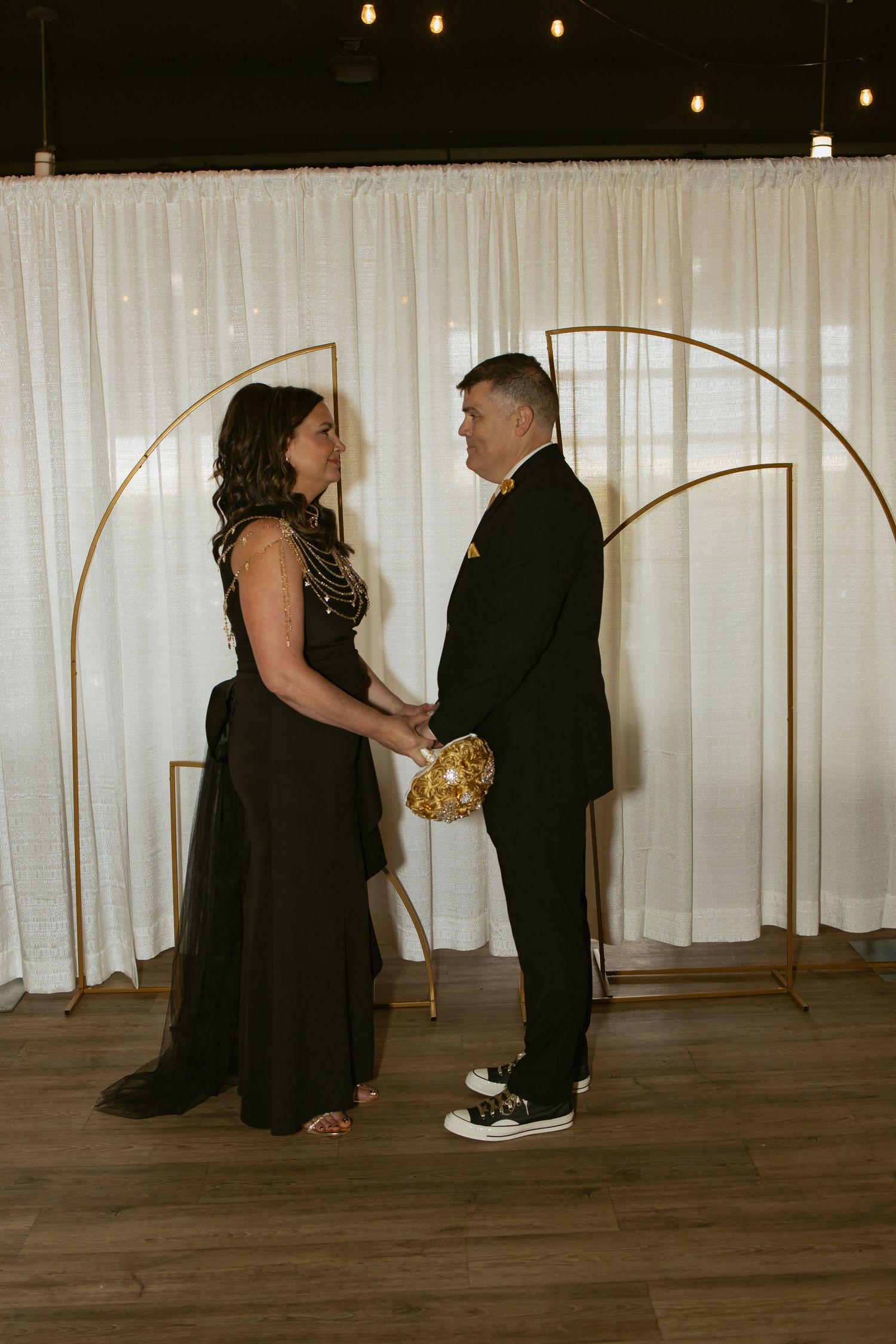 Couple in black attire holding gold bouquet during wedding ceremony in front of gold arch with white drape backdrop.