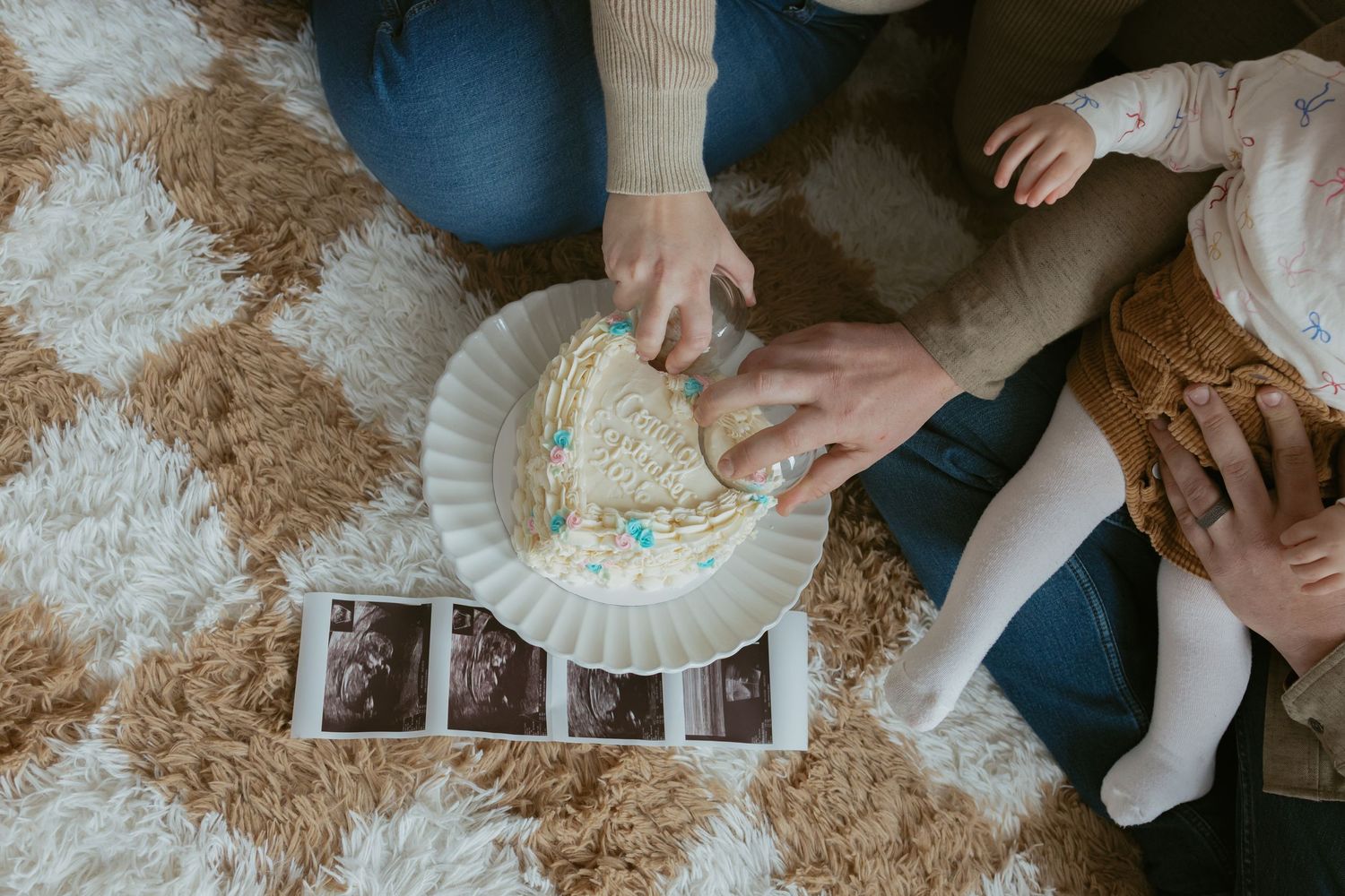 Family cutting a gender reveal cake with blue filling on a fluffy rug beside ultrasound photos.