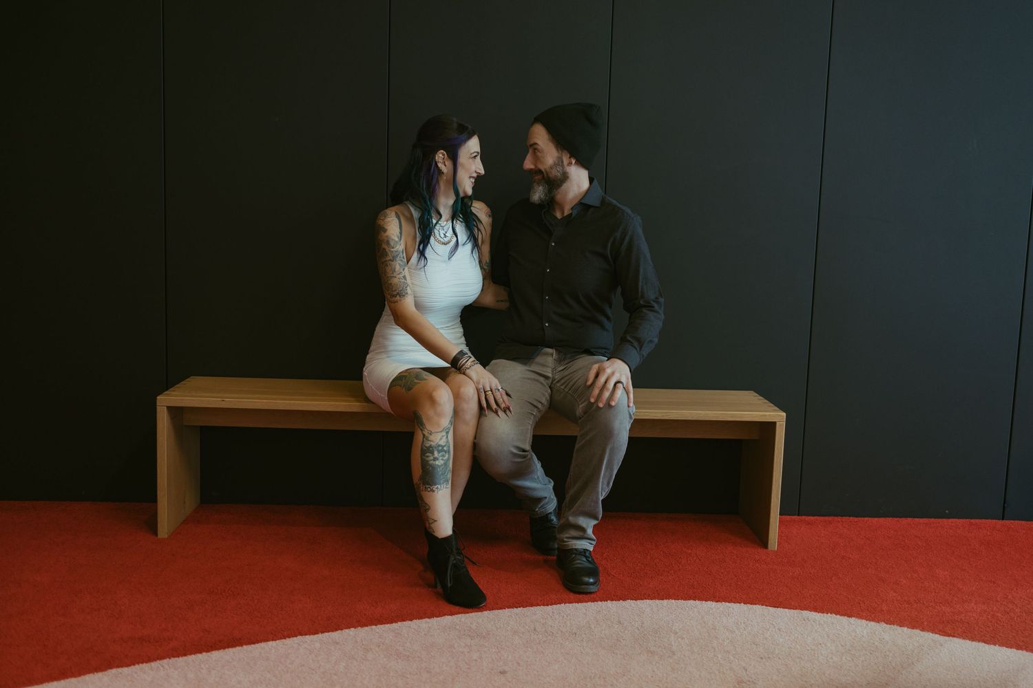 A tattooed couple sits on a wooden bench, smiling at each other on a red carpet floor.