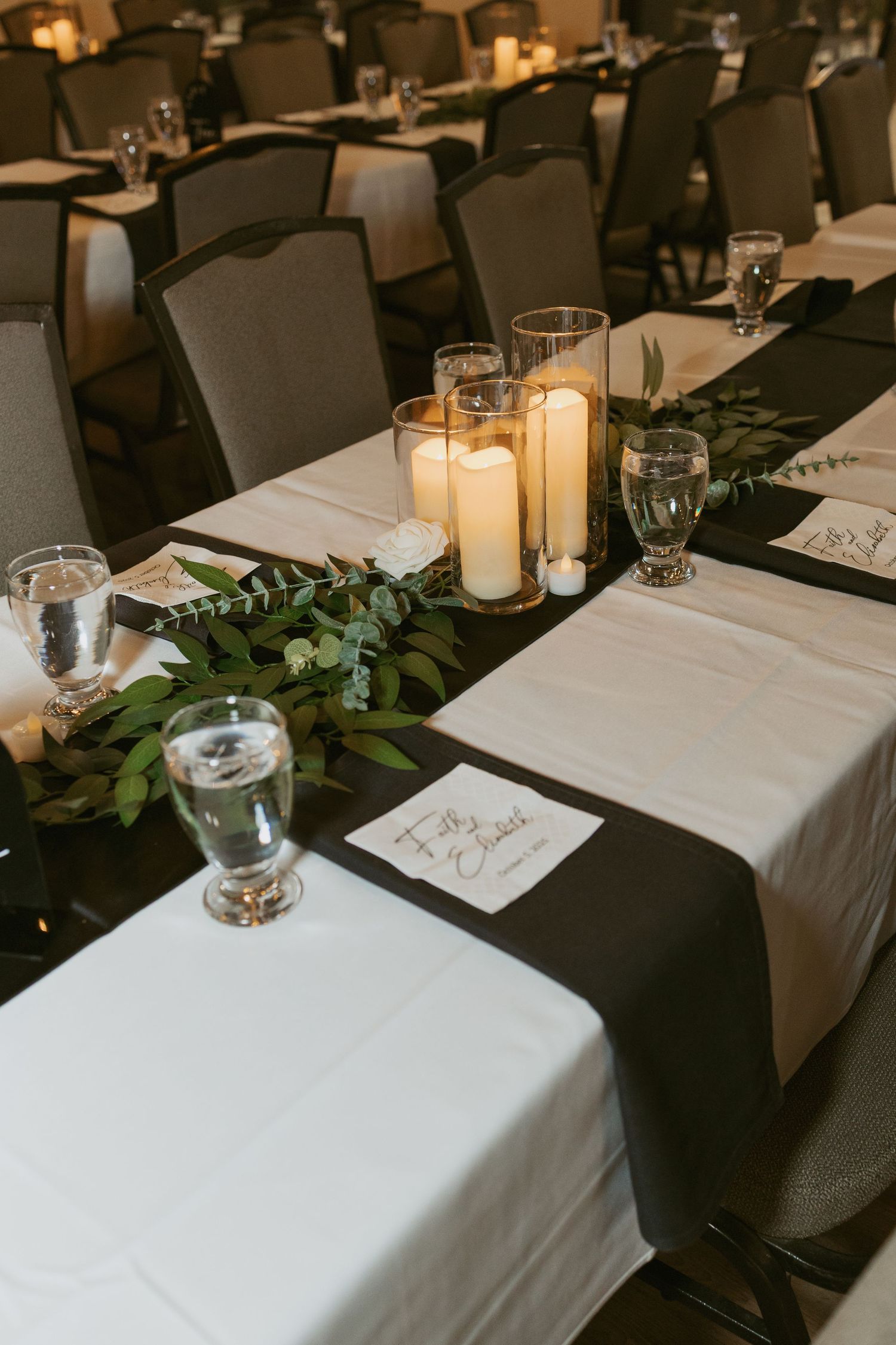 Elegant wedding reception tables with white linens, black runners, greenery garlands, and glowing pillar candles in glass holders.