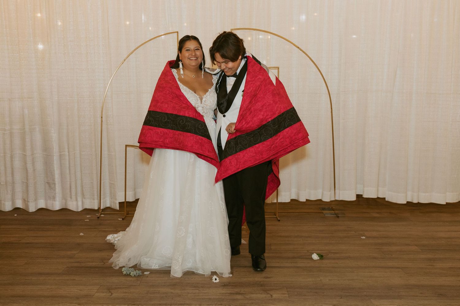 Bride and groom wrapped together in a red and black blanket, standing under a gold arch at their wedding reception.