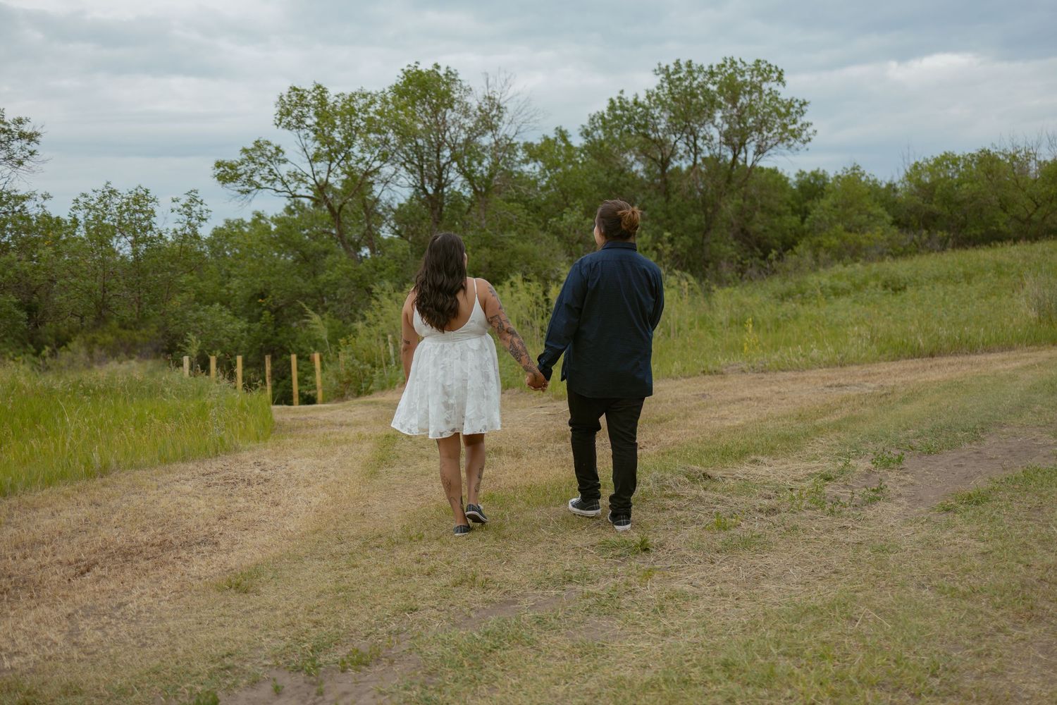 A couple walks down a dirt path surrounded by green grass and trees under cloudy skies.