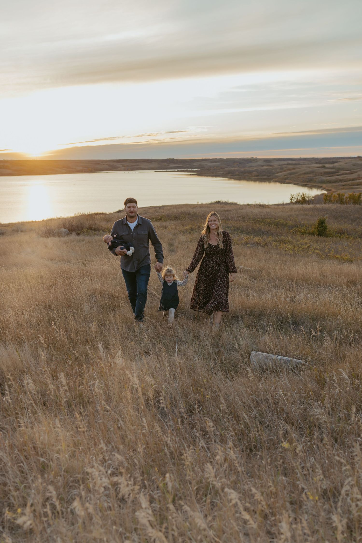 Family walking through tall prairie grass during sunset with water view in background.