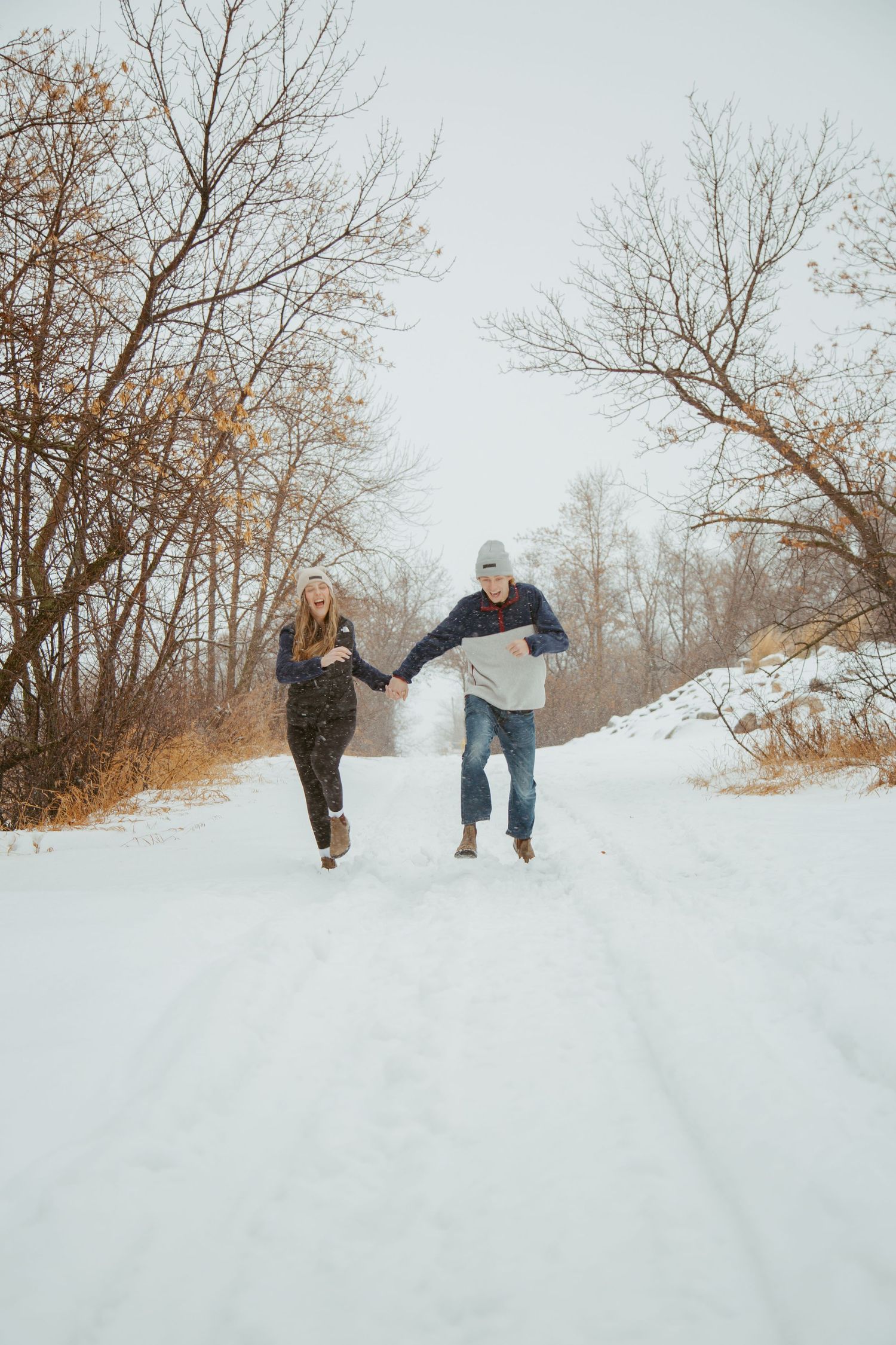 Two people running hand in hand through a snowy winter path surrounded by bare trees.