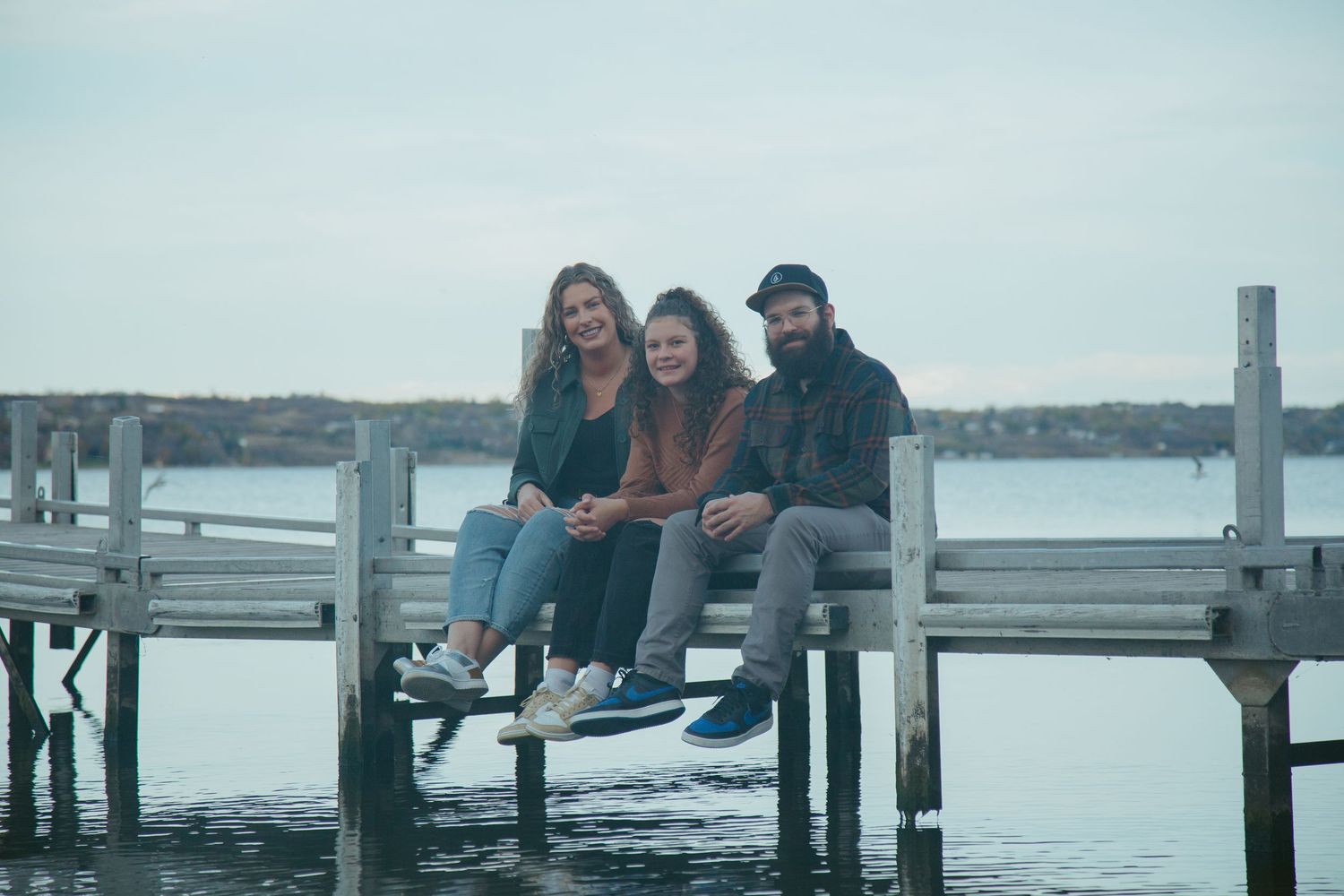 Several people sit together on a wooden dock overlooking a peaceful waterfront.