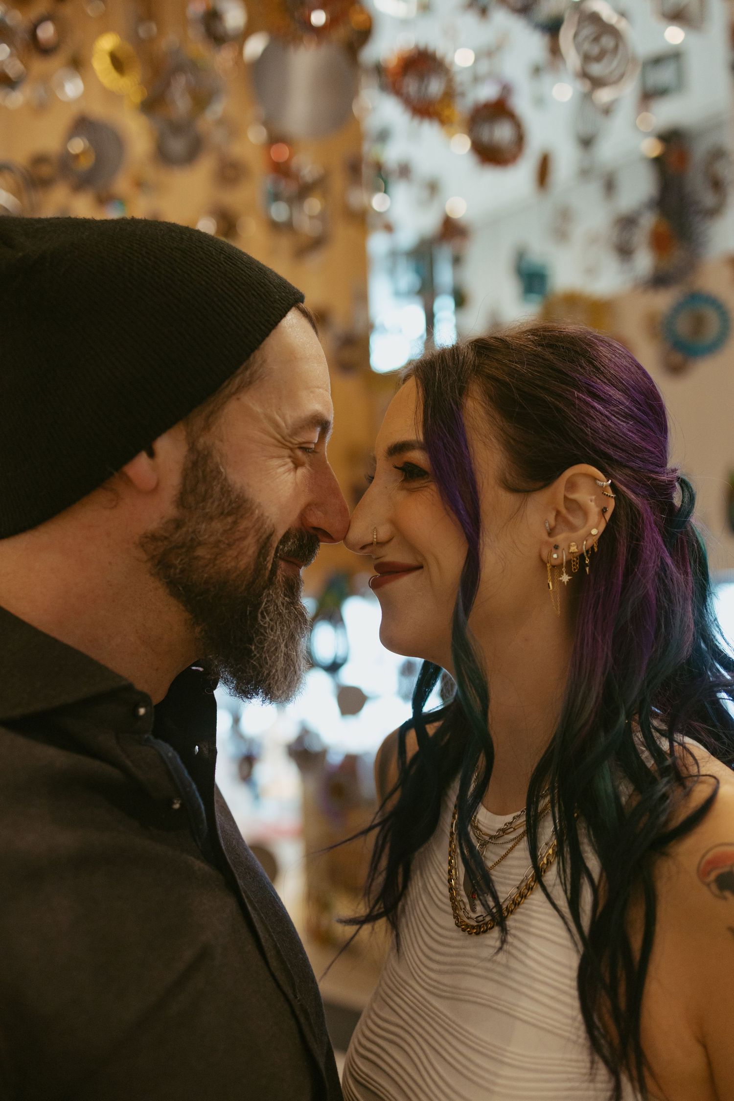 A bearded man in a black beanie and a woman with purple hair share a romantic nose-to-nose moment indoors.