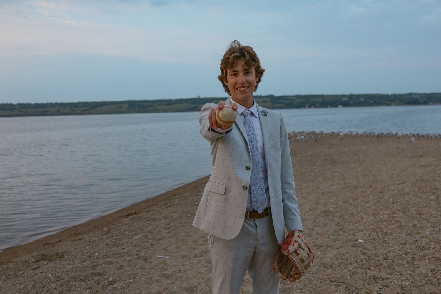 A figure in formal attire stands on a sandy beach with a waterfront view at sunset.