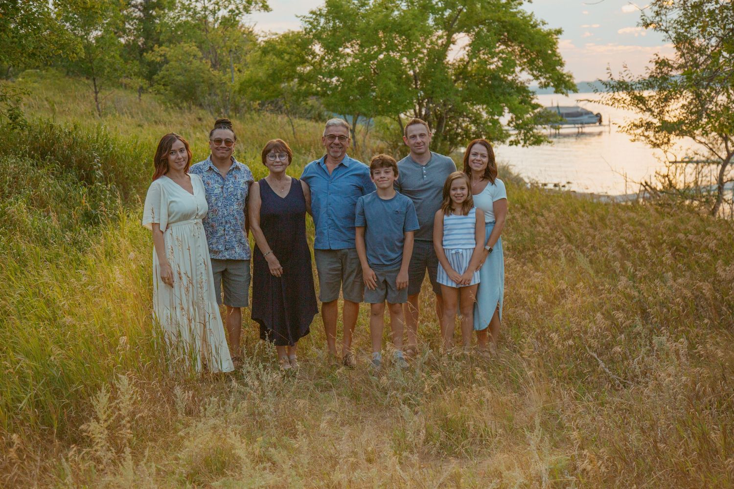 Family group poses on grassy hillside at sunset with water and boats in background.