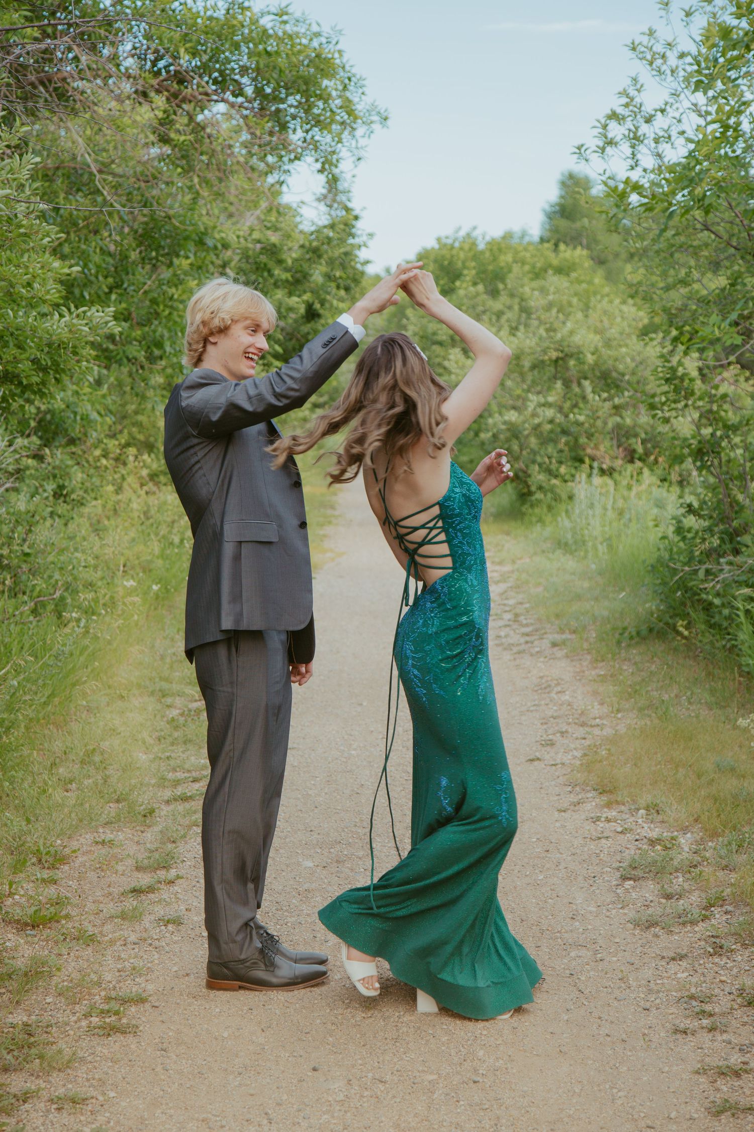 Two people dance on a nature path, one wearing a grey suit and another in an emerald green dress.