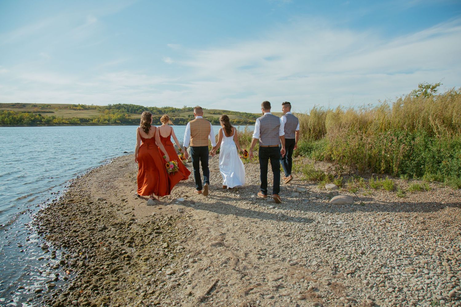 Wedding party walks along a rocky shoreline with blue water and sky in the background.