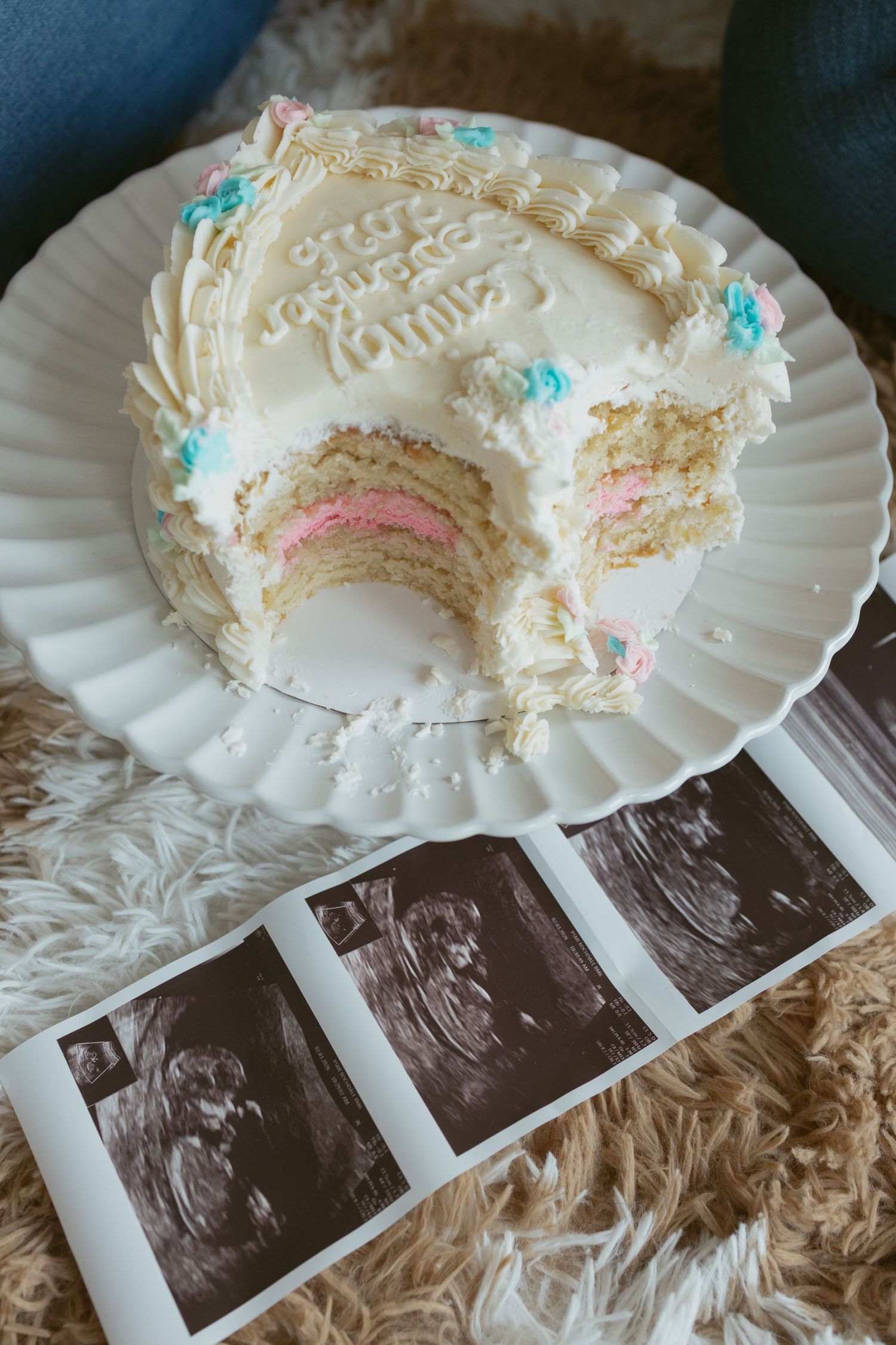 Gender reveal cake with pink layers beside ultrasound photos on a fluffy rug, decorated with blue and pink frosting.