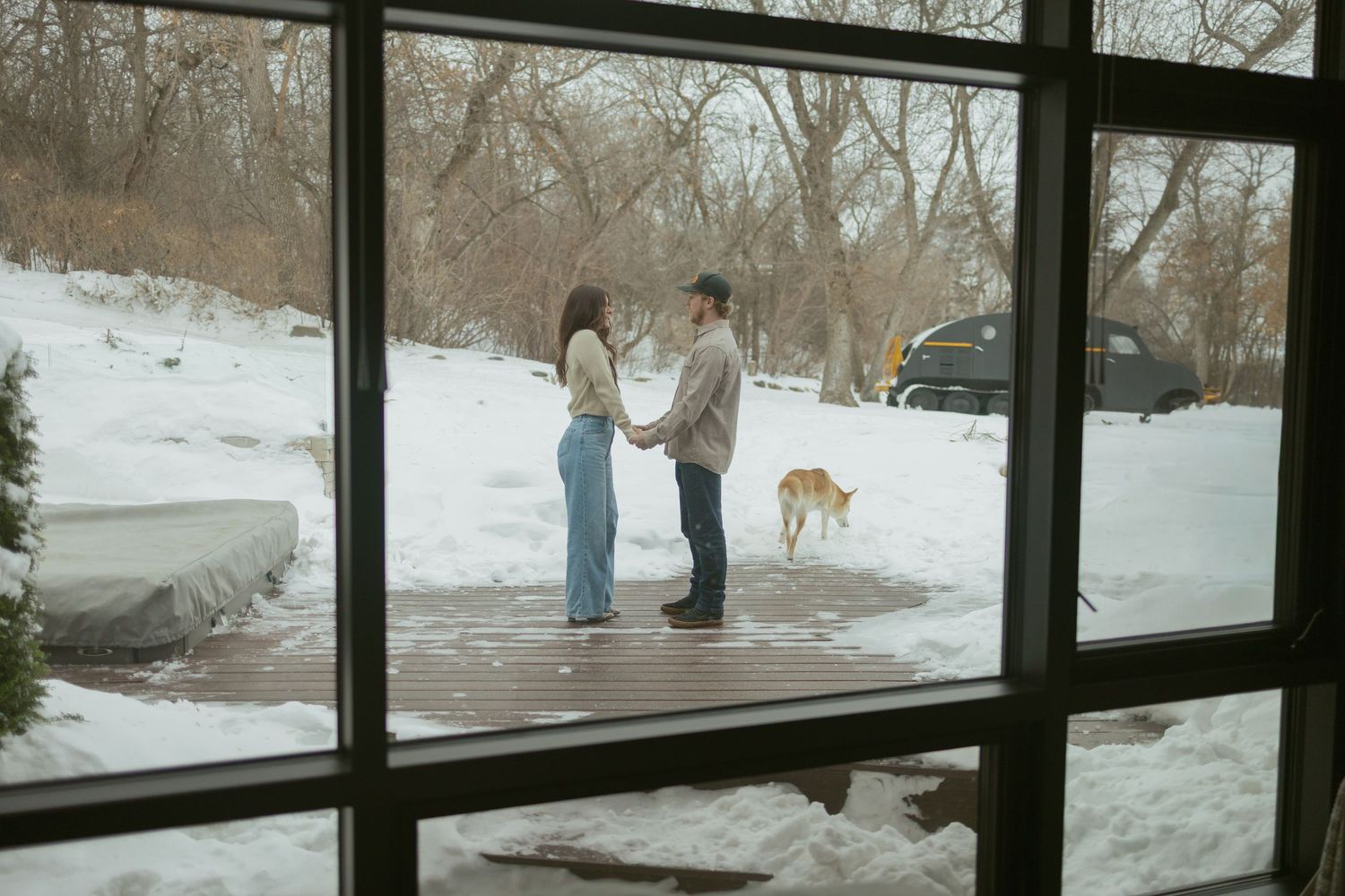Couple holding hands in snowy outdoor setting viewed through large window with dog in background.