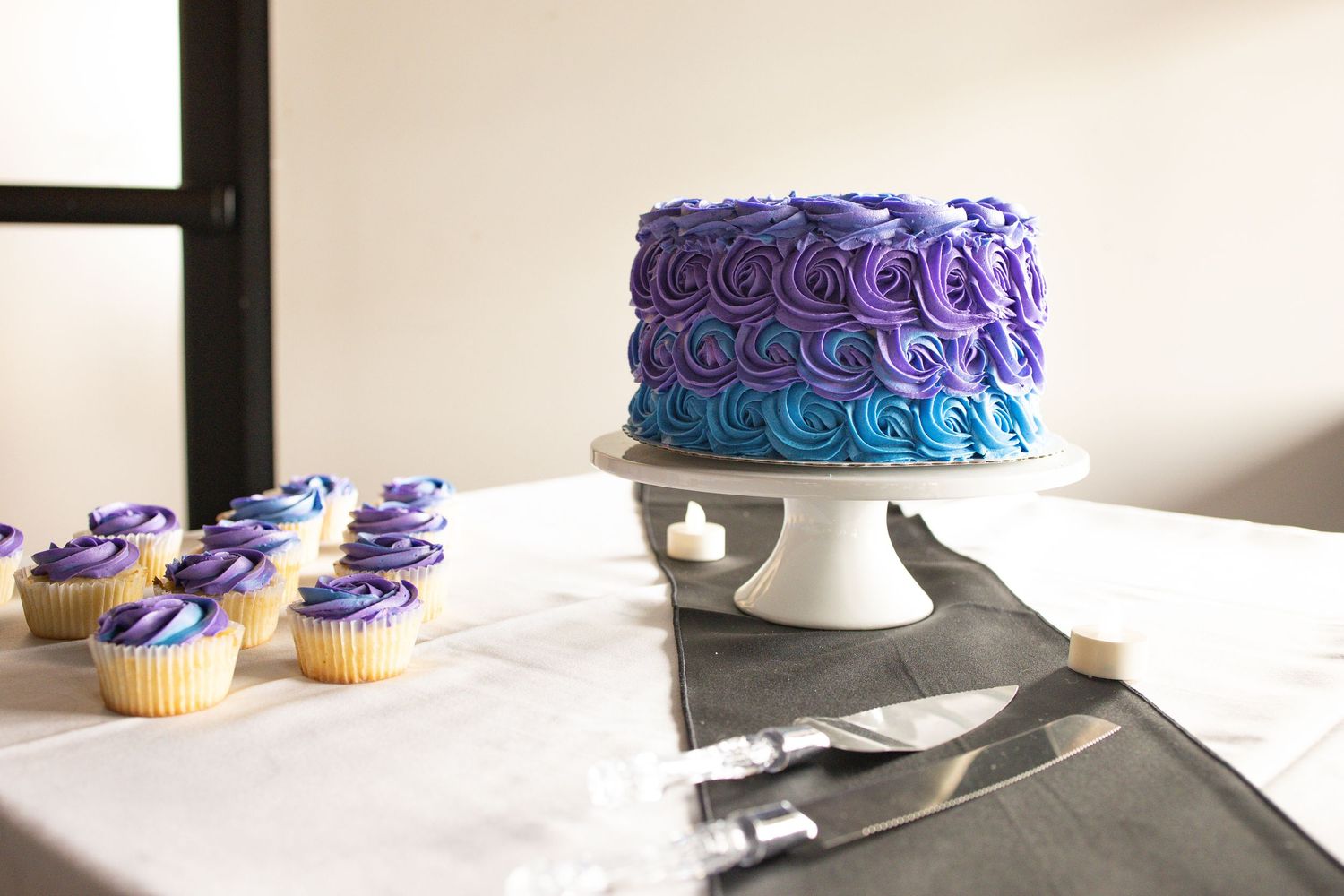 Purple and blue ombre rosette cake on white stand with matching cupcakes and serving knife on white tablecloth.
