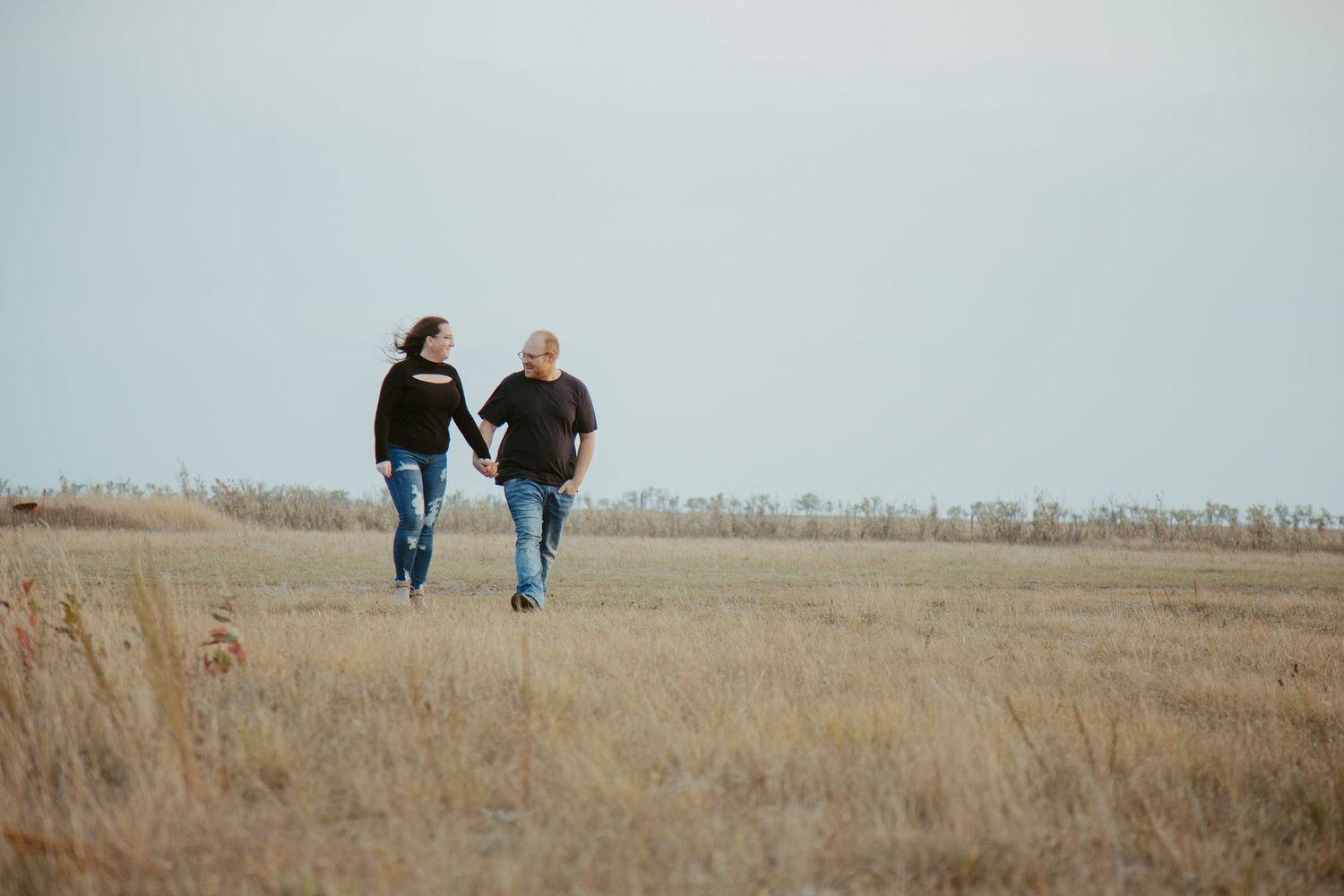 Couple holding hands walking through golden prairie field under overcast sky.