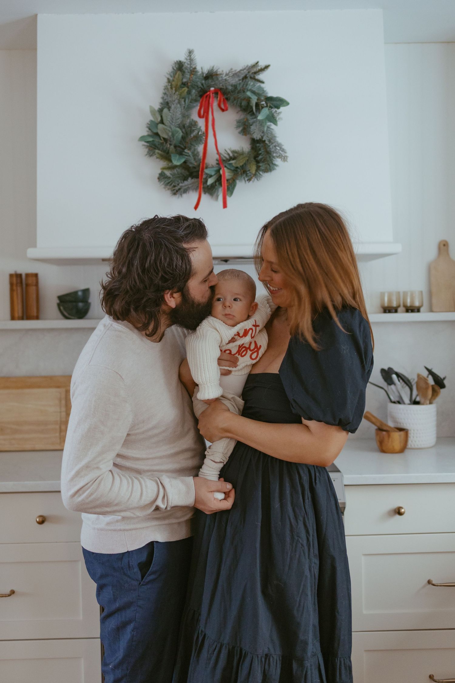 Family shares intimate holiday moments in a modern white kitchen decorated with Christmas wreaths.
