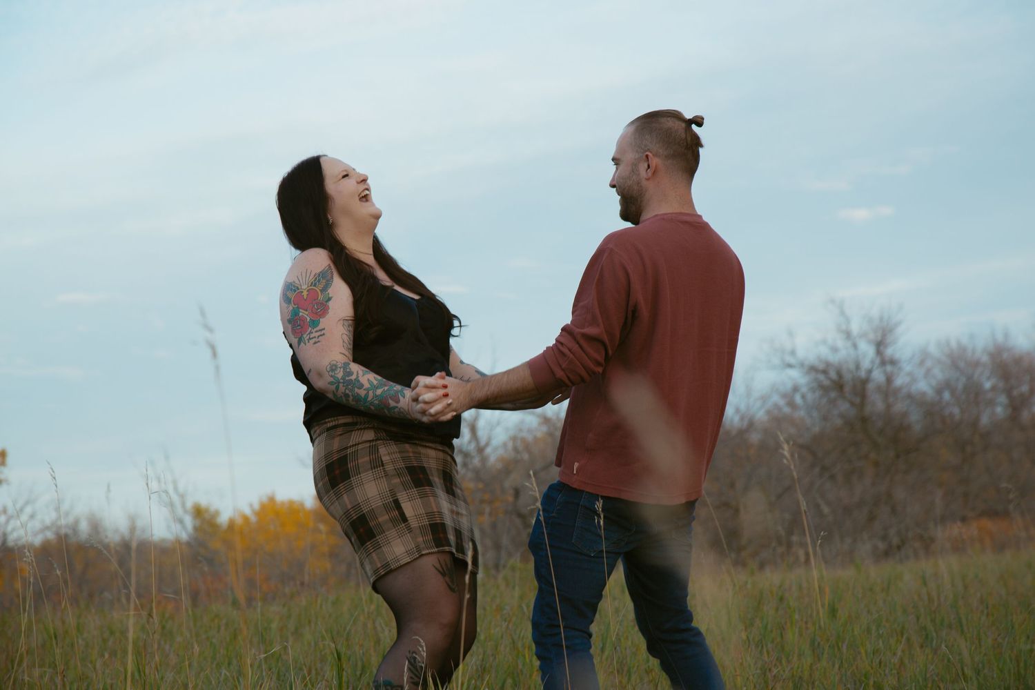 A couple laughs together while dancing in a grassy field during autumn.