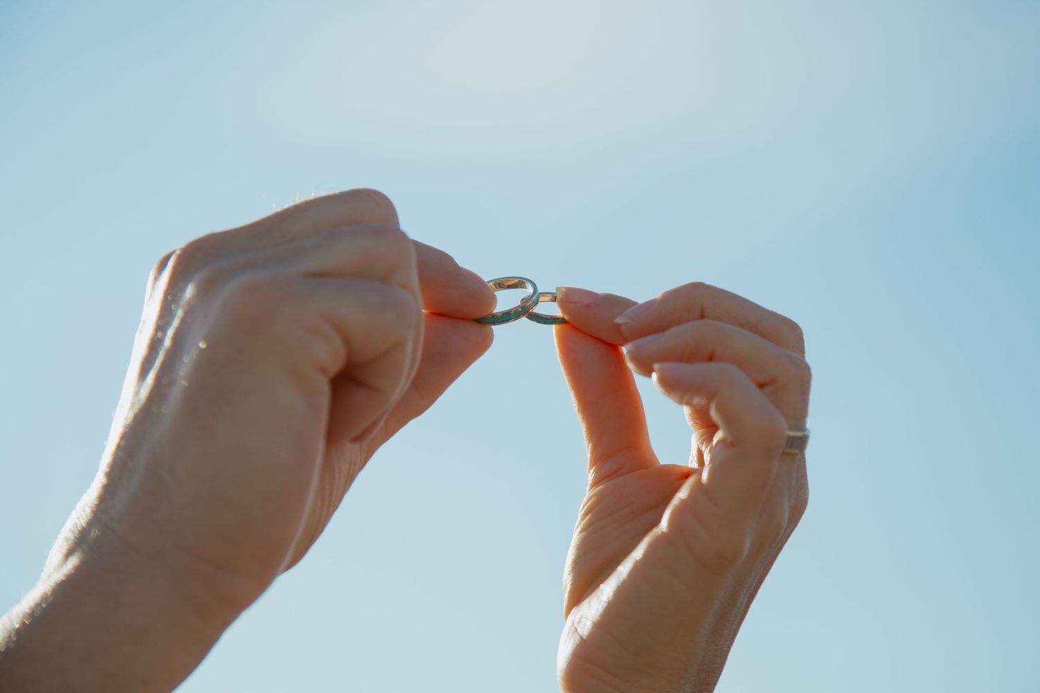 Hands holding a wedding ring against bright blue sky background.