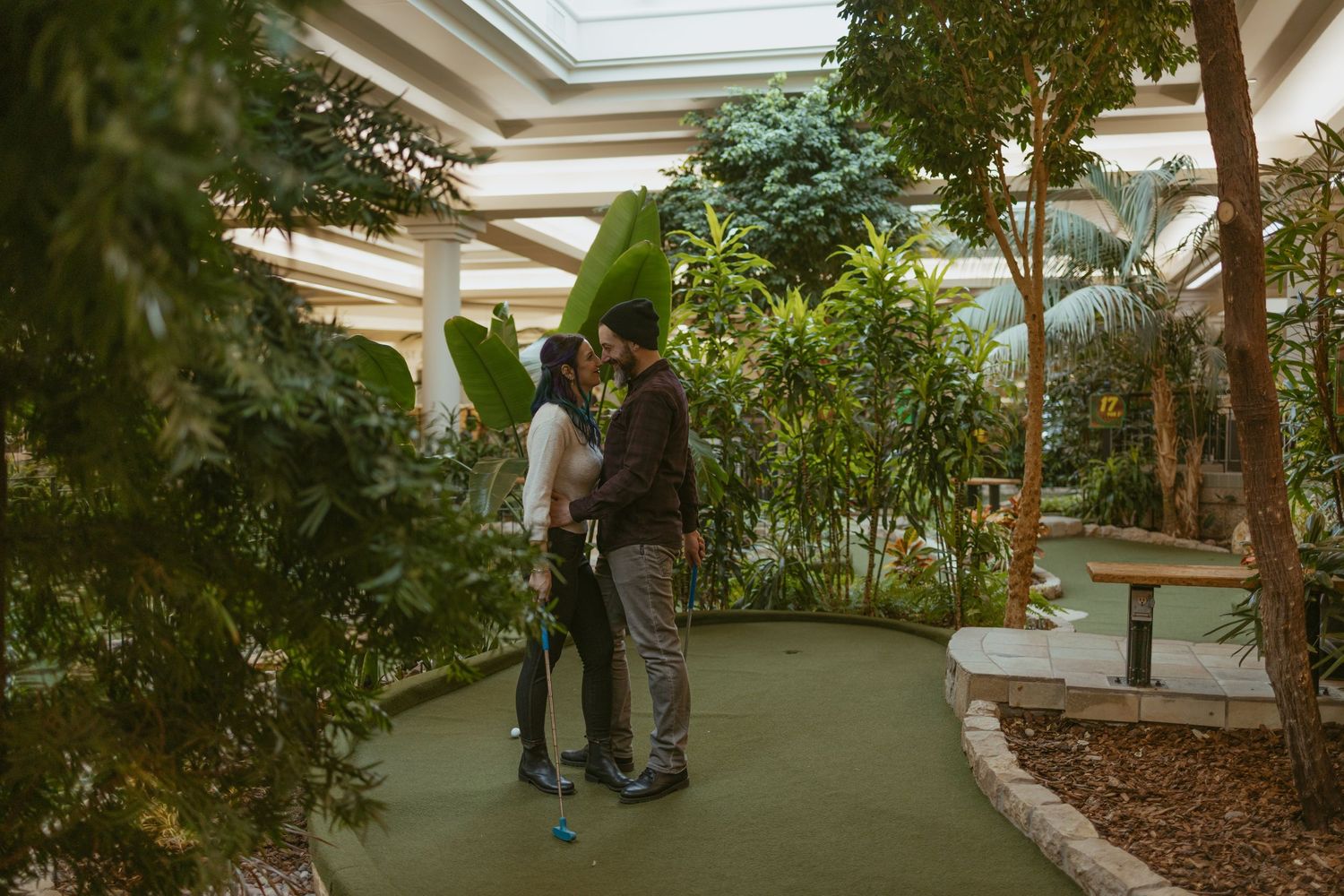 Couple sharing a romantic kiss on an indoor mini golf course surrounded by lush tropical plants.