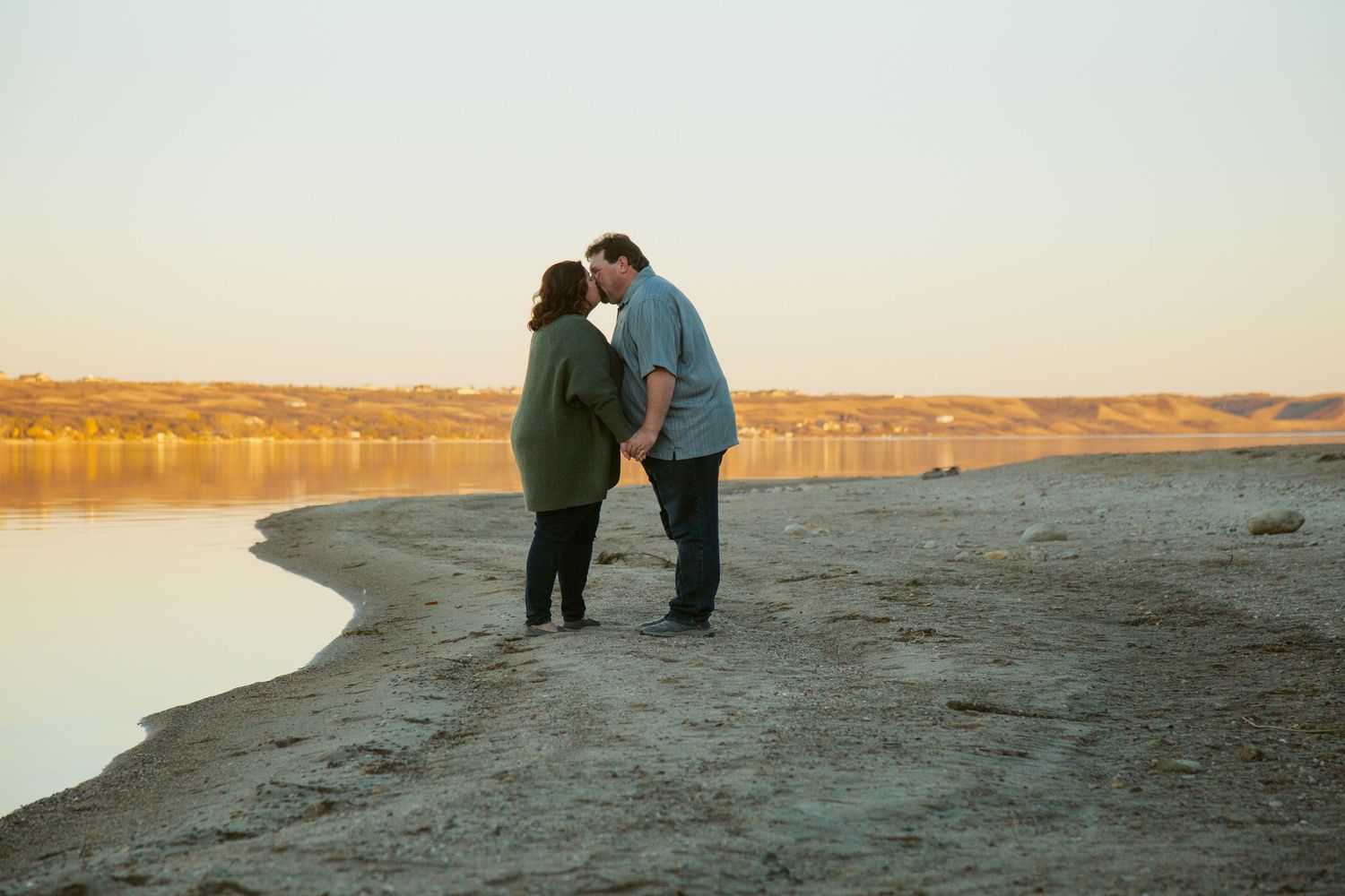 Couple walking along a shoreline during sunset with water reflecting golden light.