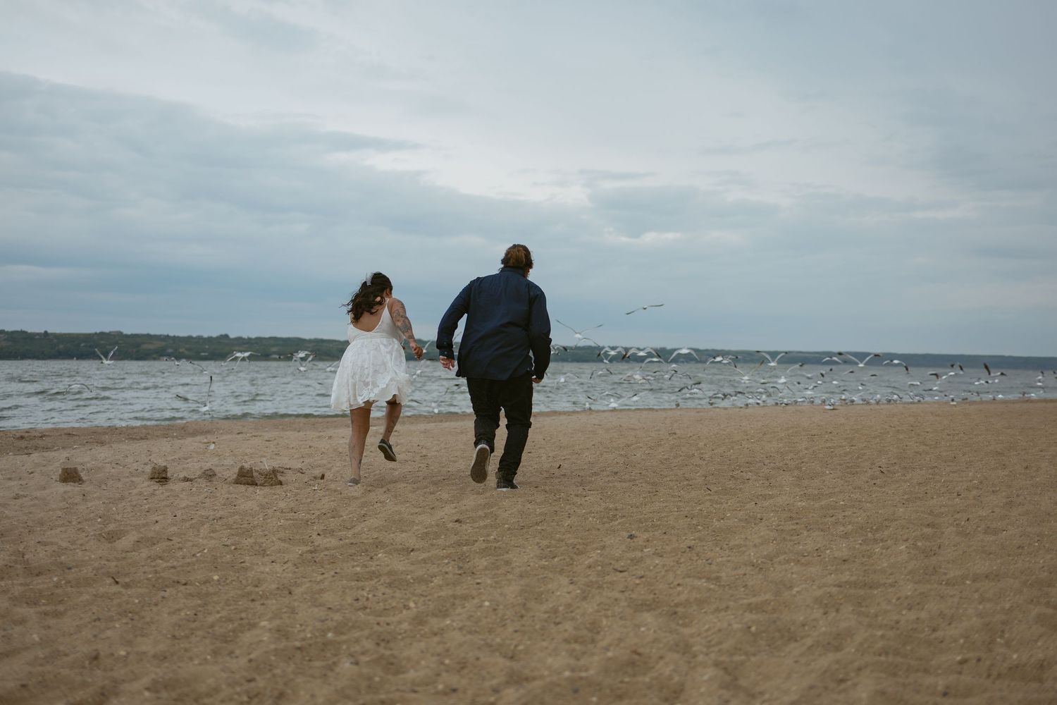 A couple walks together on a beach with waves and cloudy skies in the background.