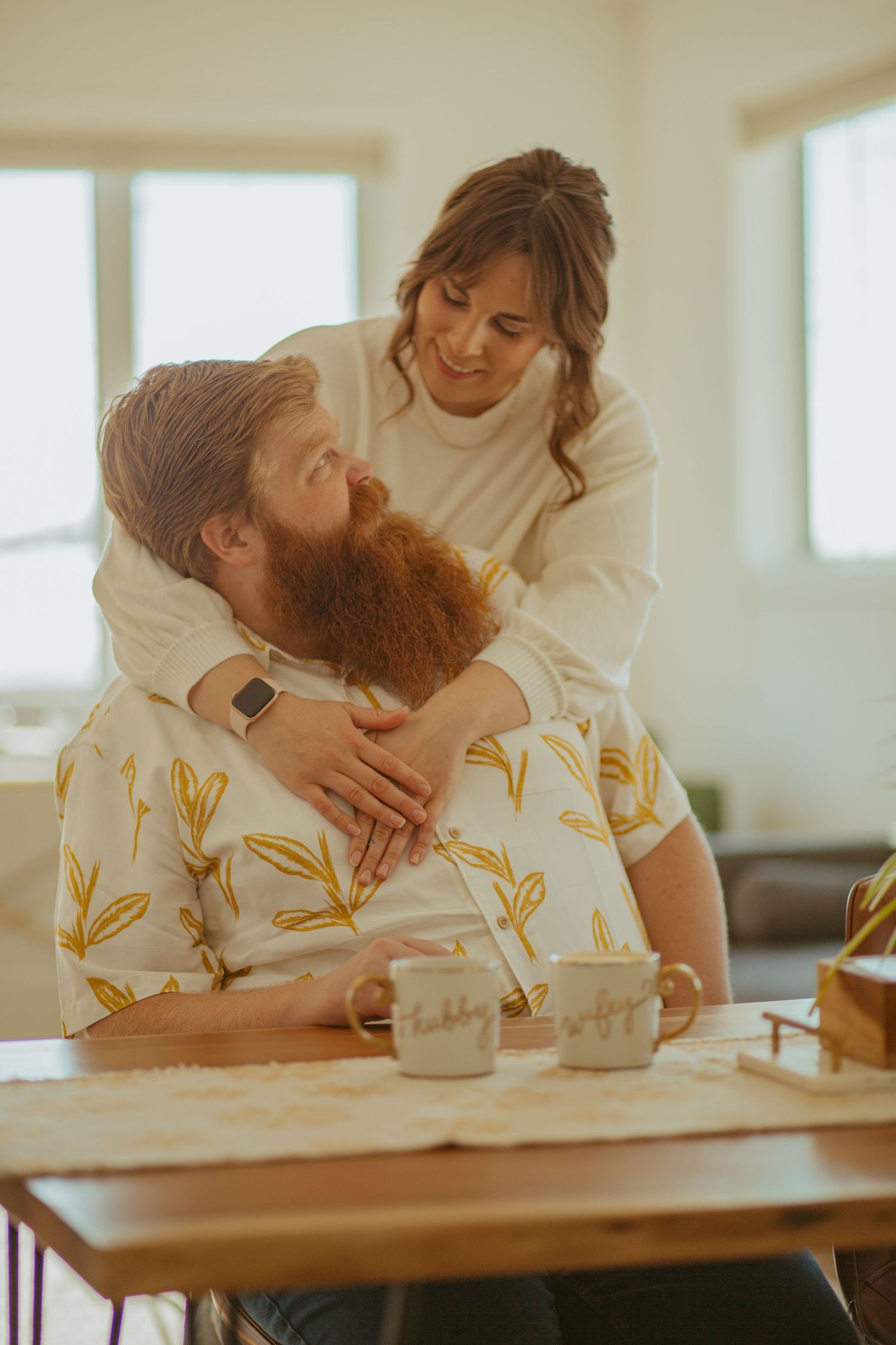 Couple embracing in casual attire near a wooden table in a sunlit room.