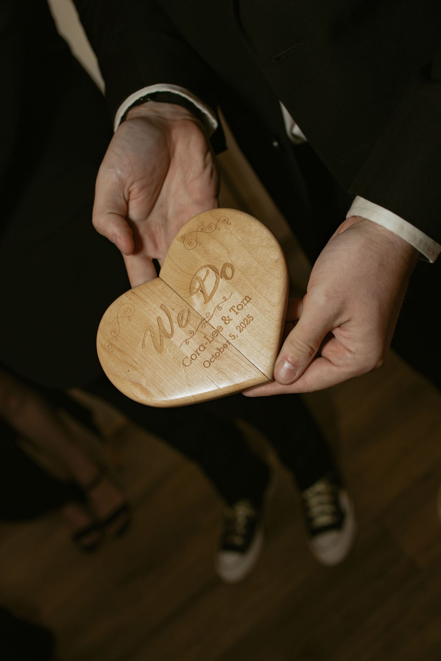 Person in a suit holding a heart-shaped wooden keepsake engraved with We Do, featuring a couple's names and wedding date.