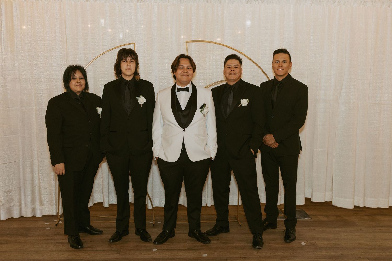Five groomsmen in black tuxedos pose together, with the groom in a white jacket, against a draped white backdrop.