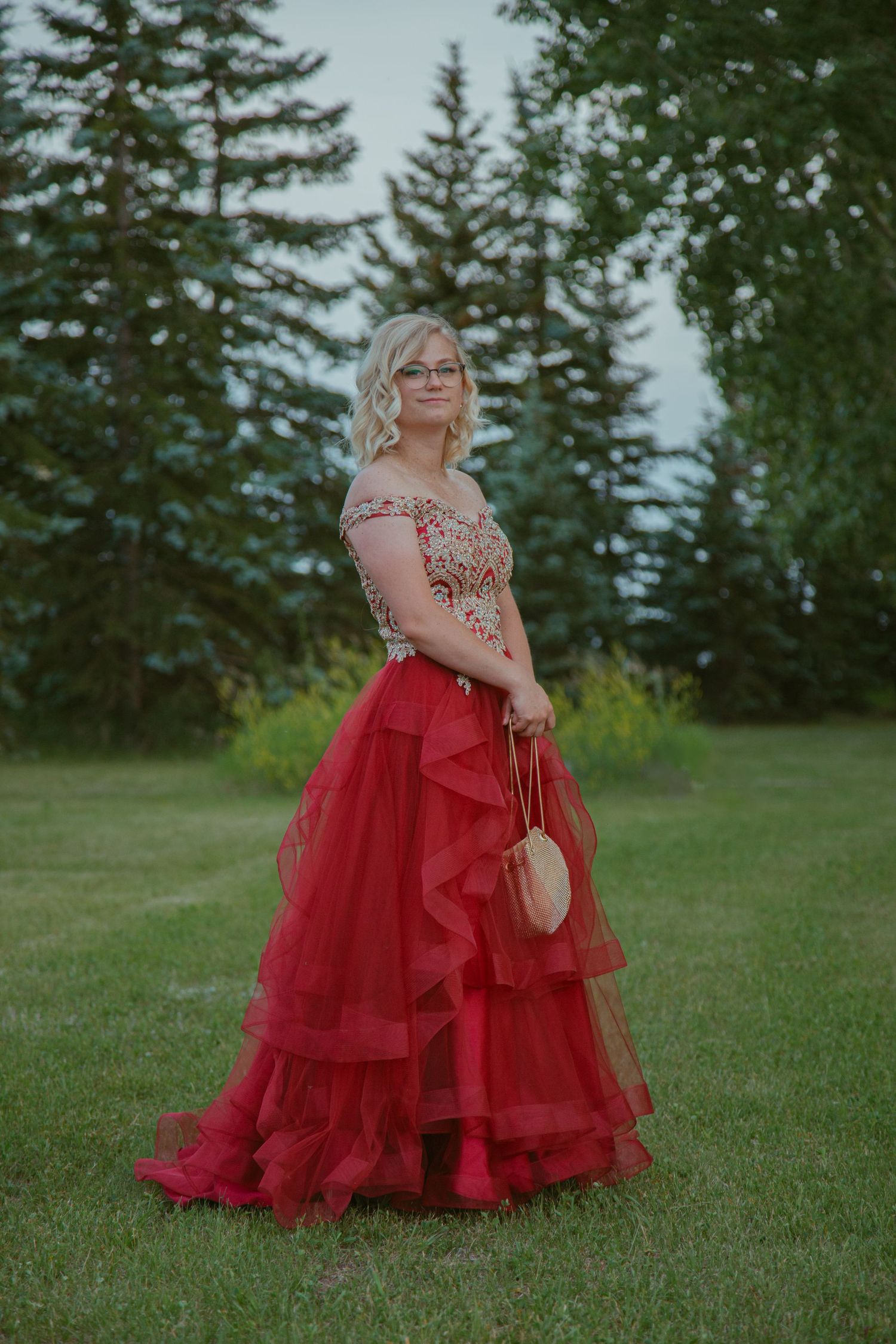 Elegant red formal gown photographed in outdoor setting with evergreen trees.