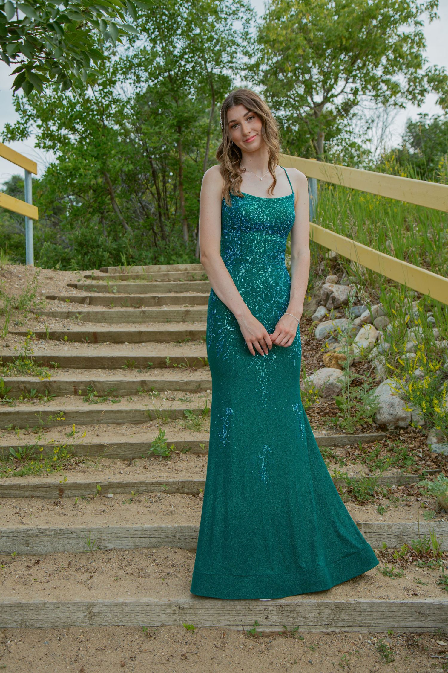 A person in a teal summer dress poses on rustic outdoor stone steps surrounded by greenery.