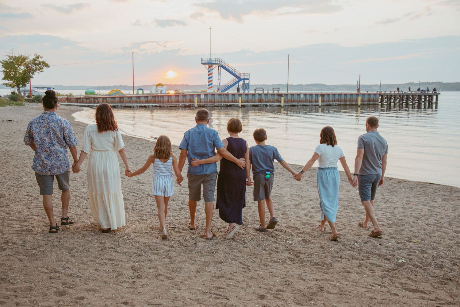 Group walking hand in hand along beach shore with pier visible in background at sunset.