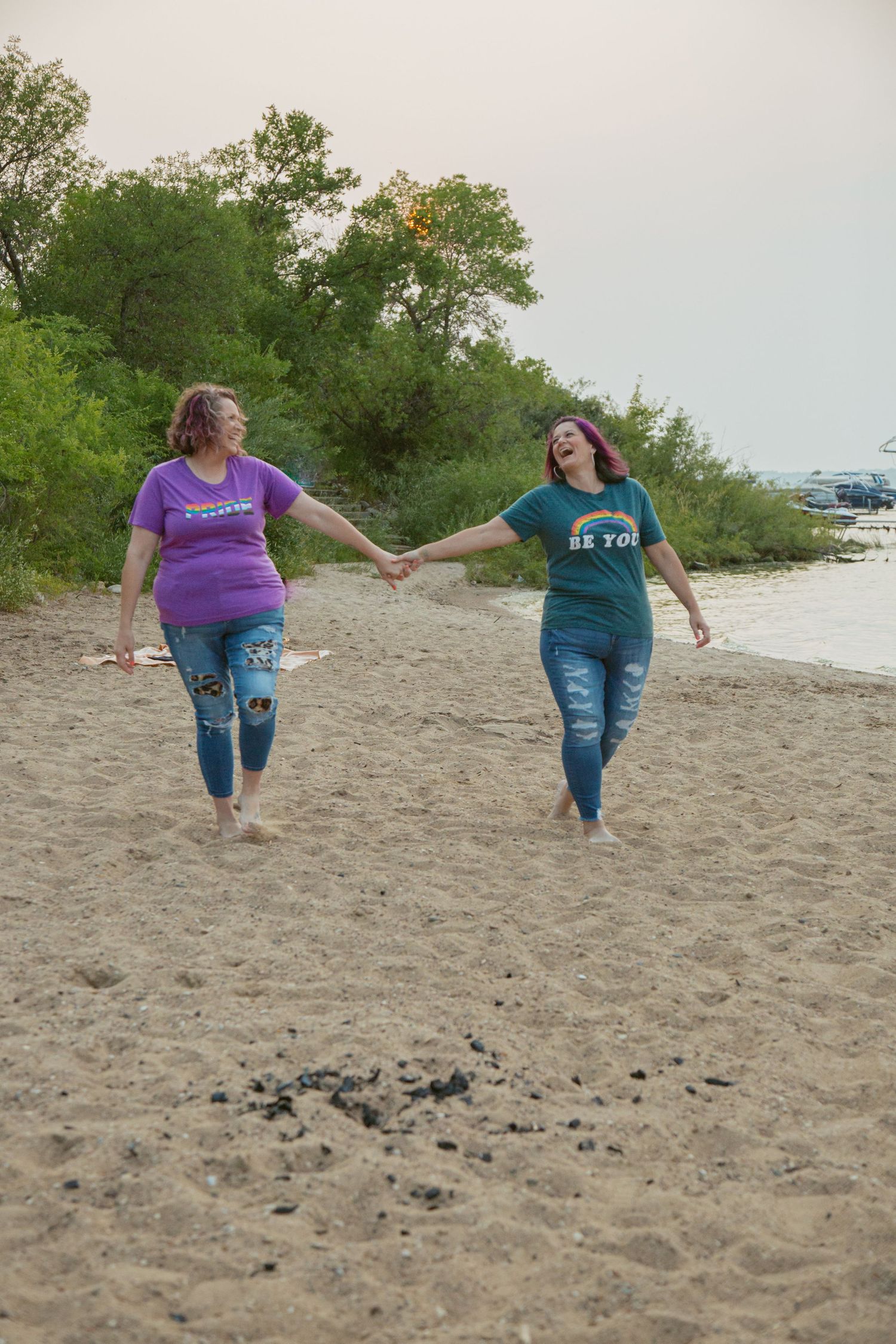 Two friends in casual wear holding hands while walking on sandy beach.