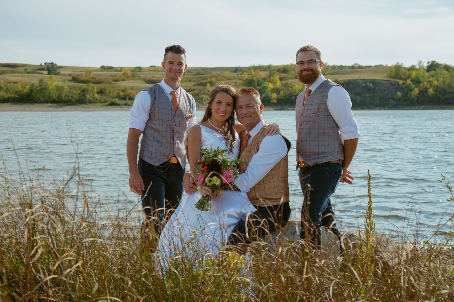 Wedding party poses together by a scenic lake during golden hour celebration.