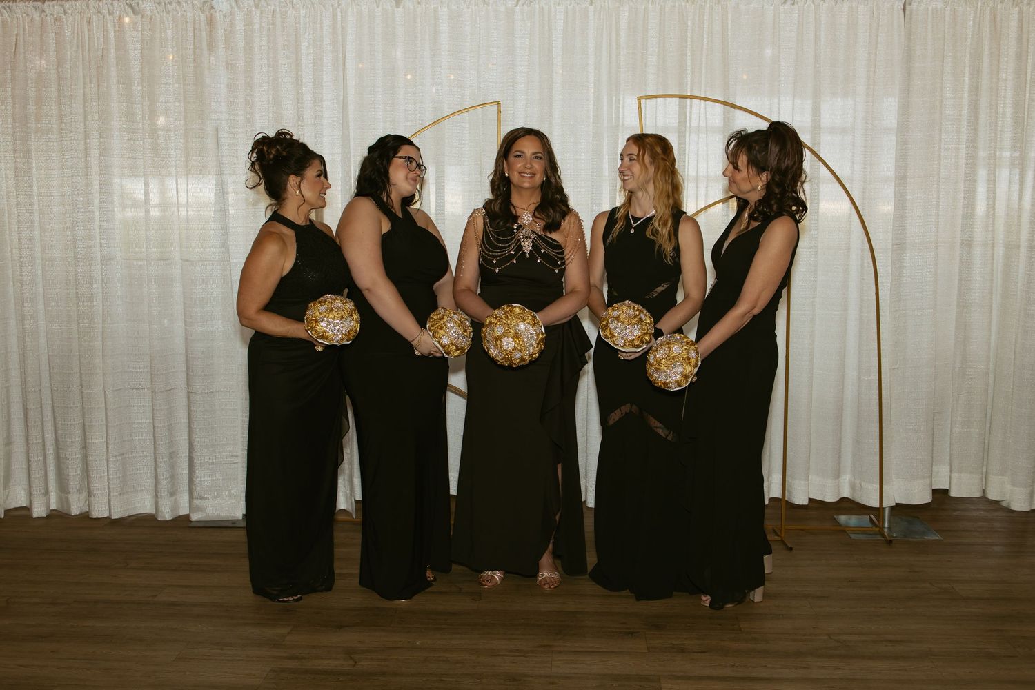 Five bridesmaids in black gowns holding gold bouquets pose under golden arch backdrop with white curtains.