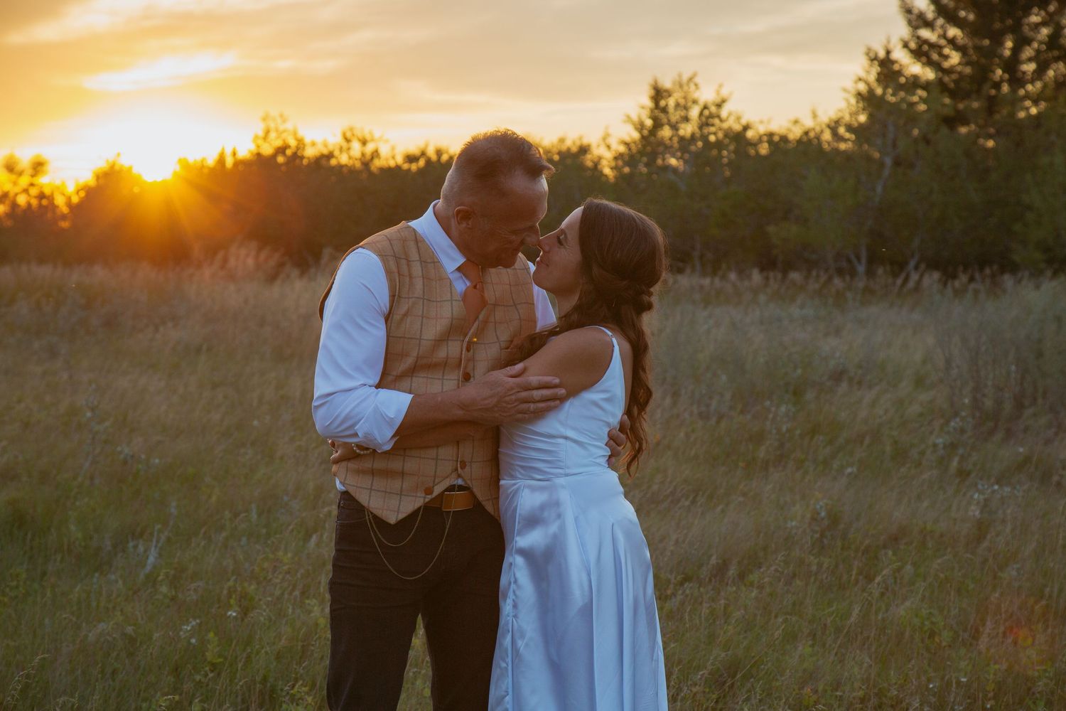 Romantic couple embraces in a golden field at sunset with sun flare and tall grass in the background.