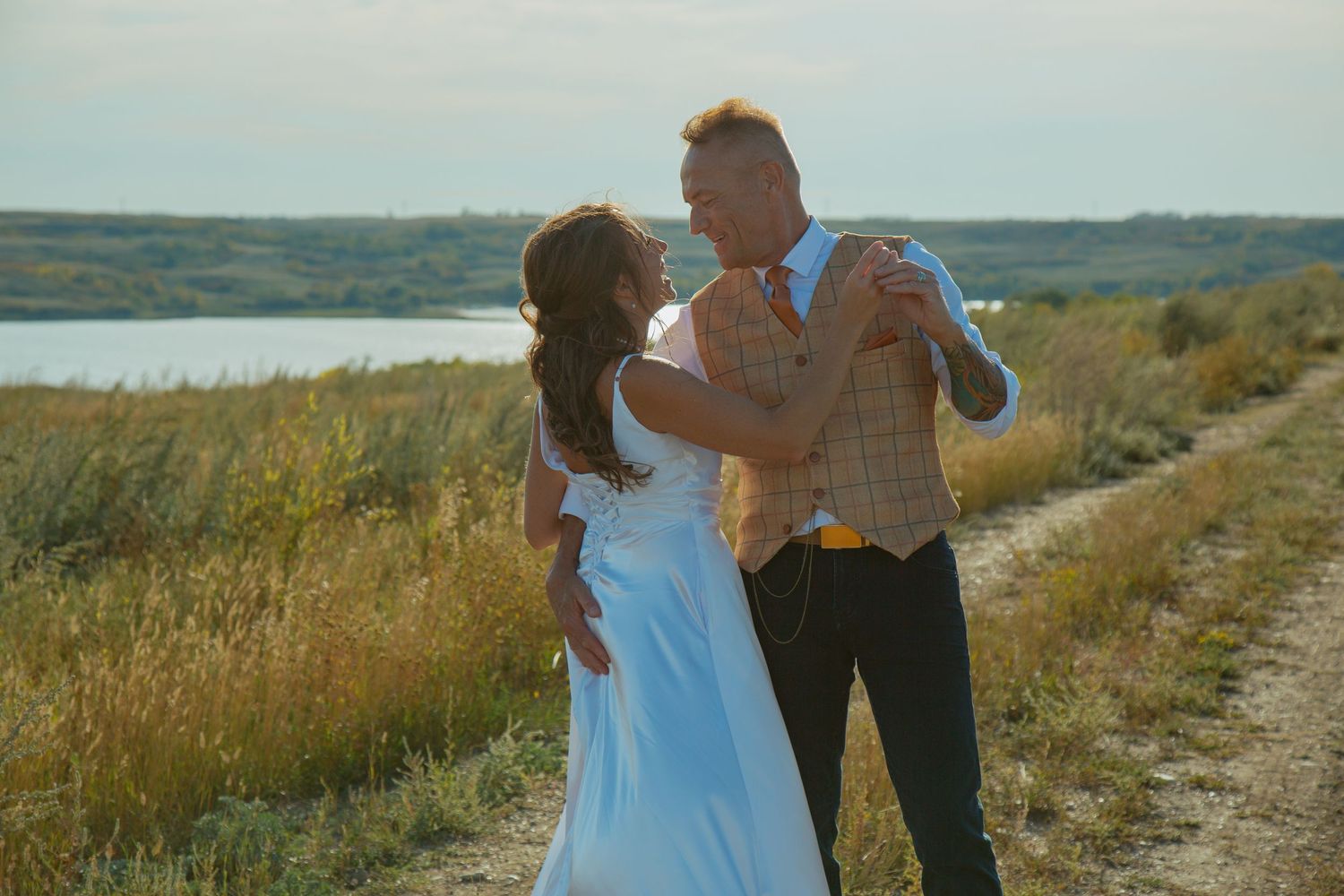 Dancing couple in wedding attire on path near lake during golden hour.