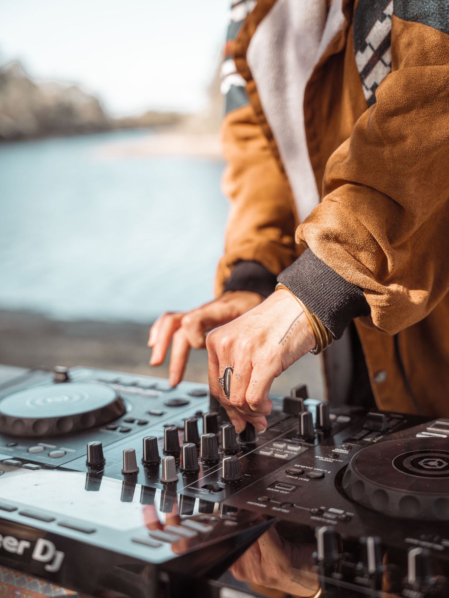 DJ mixing music outdoors on a Pioneer DJ controller near a waterfront, wearing a brown jacket and wristband.