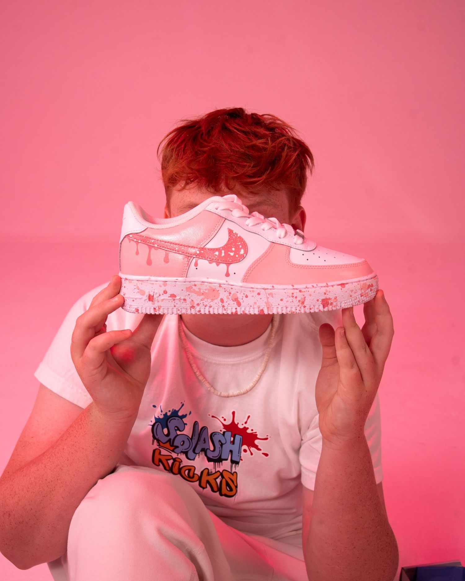 Person holding pink custom Nike Air Force 1 sneakers with drip design, wearing matching graphic tee, against pink background.