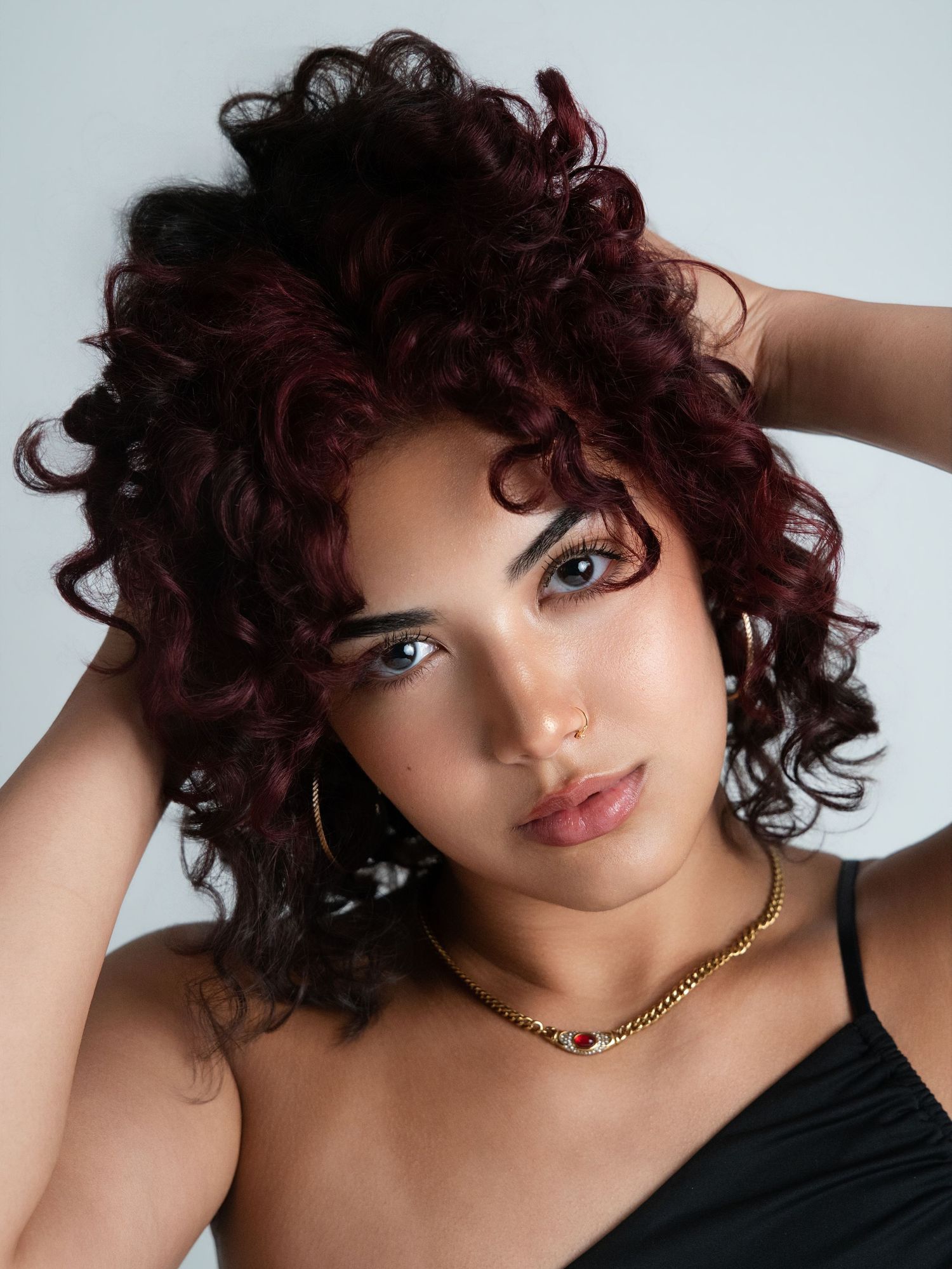 Young woman with curly dark red hair, gold chain necklace, and hoop earrings poses in a black top.