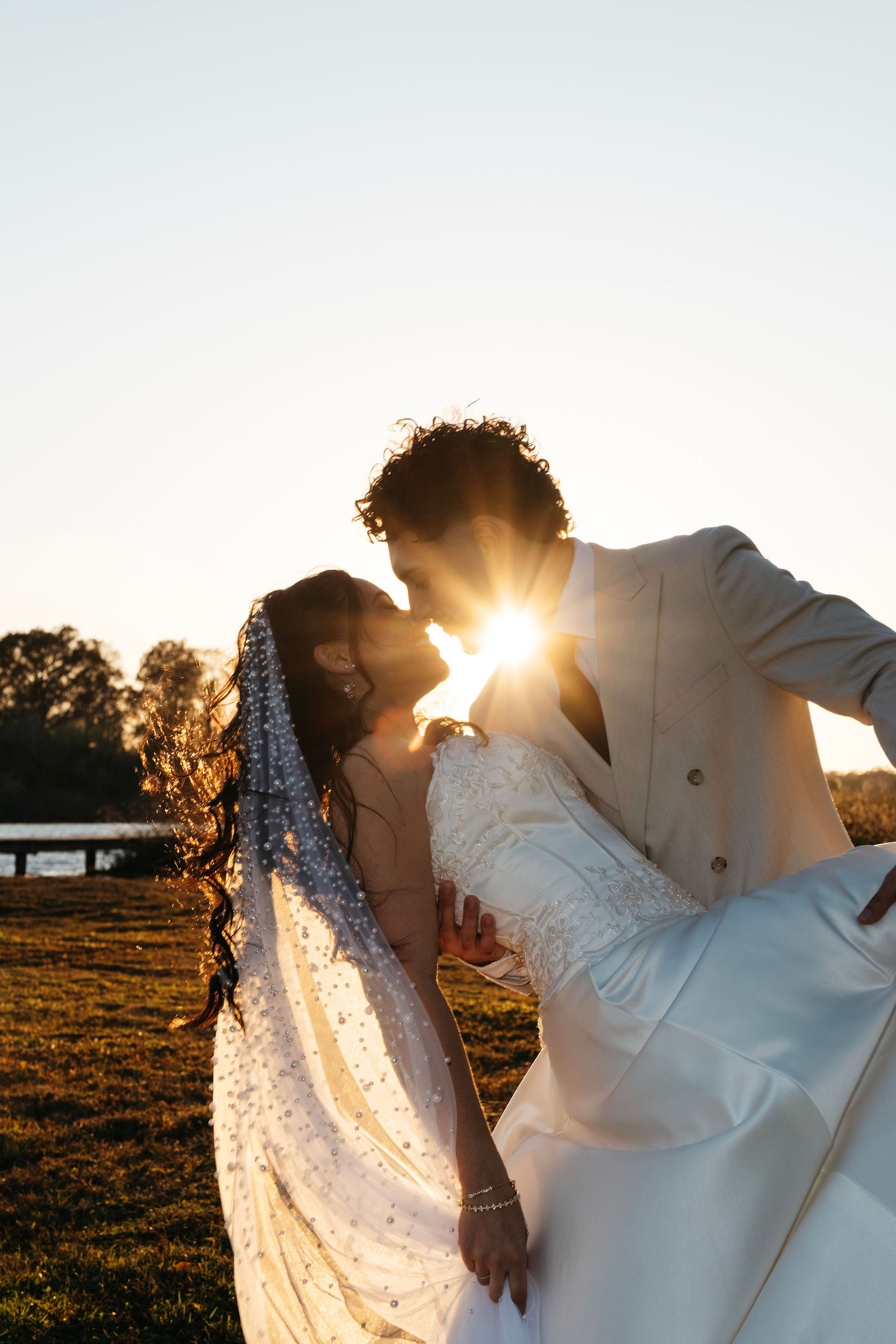 A bride and groom share a romantic sunset kiss outdoors, with golden backlight creating a stunning starburst effect.