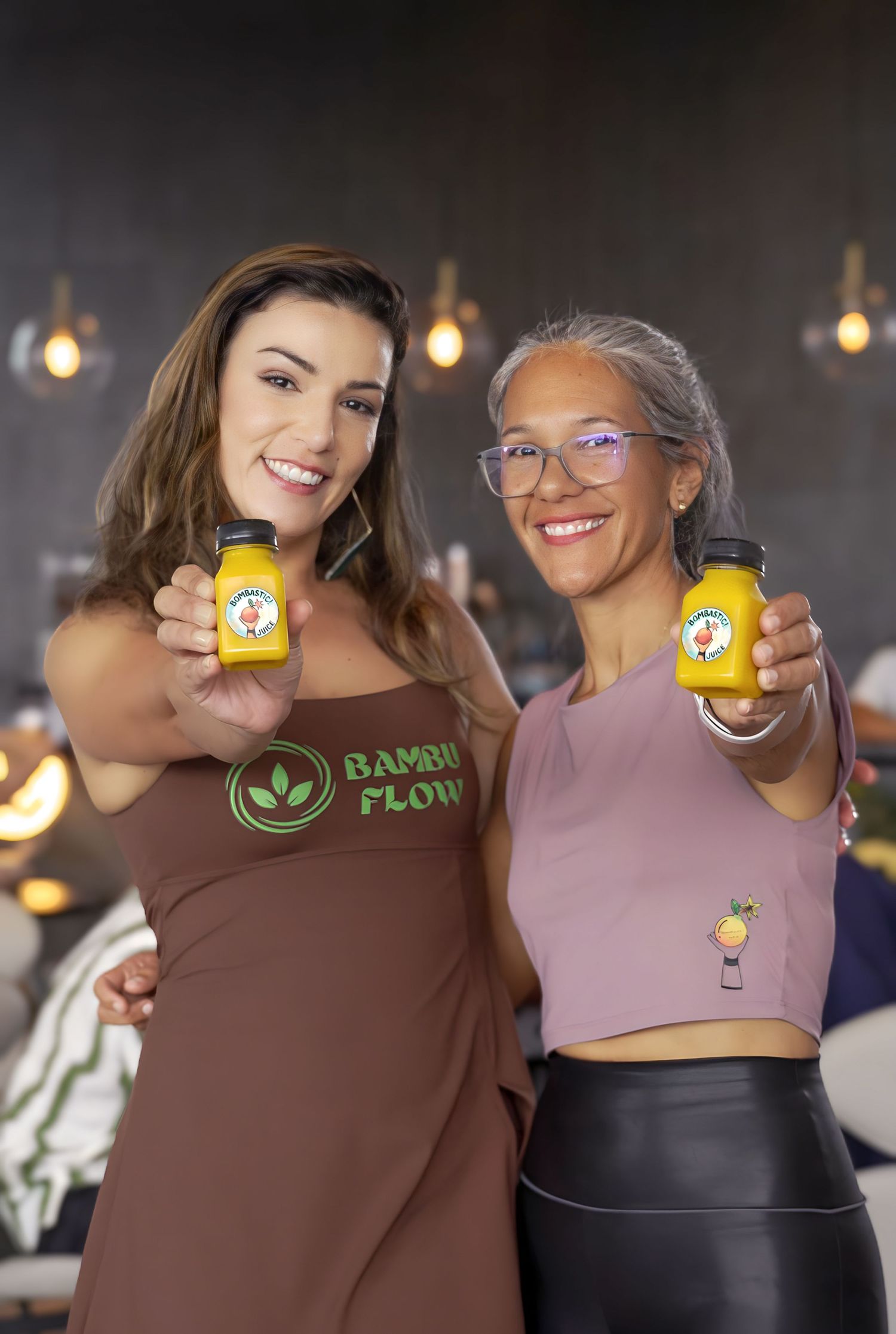 Two women smiling and holding small yellow juice bottles for Bambu Flow brand in a modern restaurant setting.