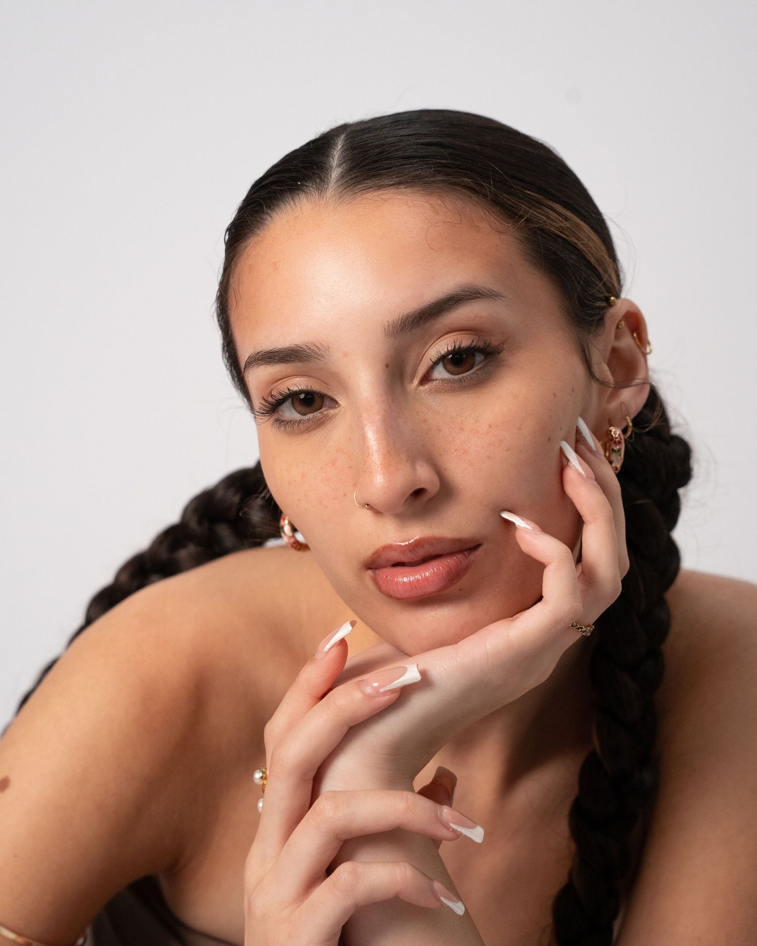 Young woman with freckles, braided hair, gold earrings, and white French tip nails poses against white background.