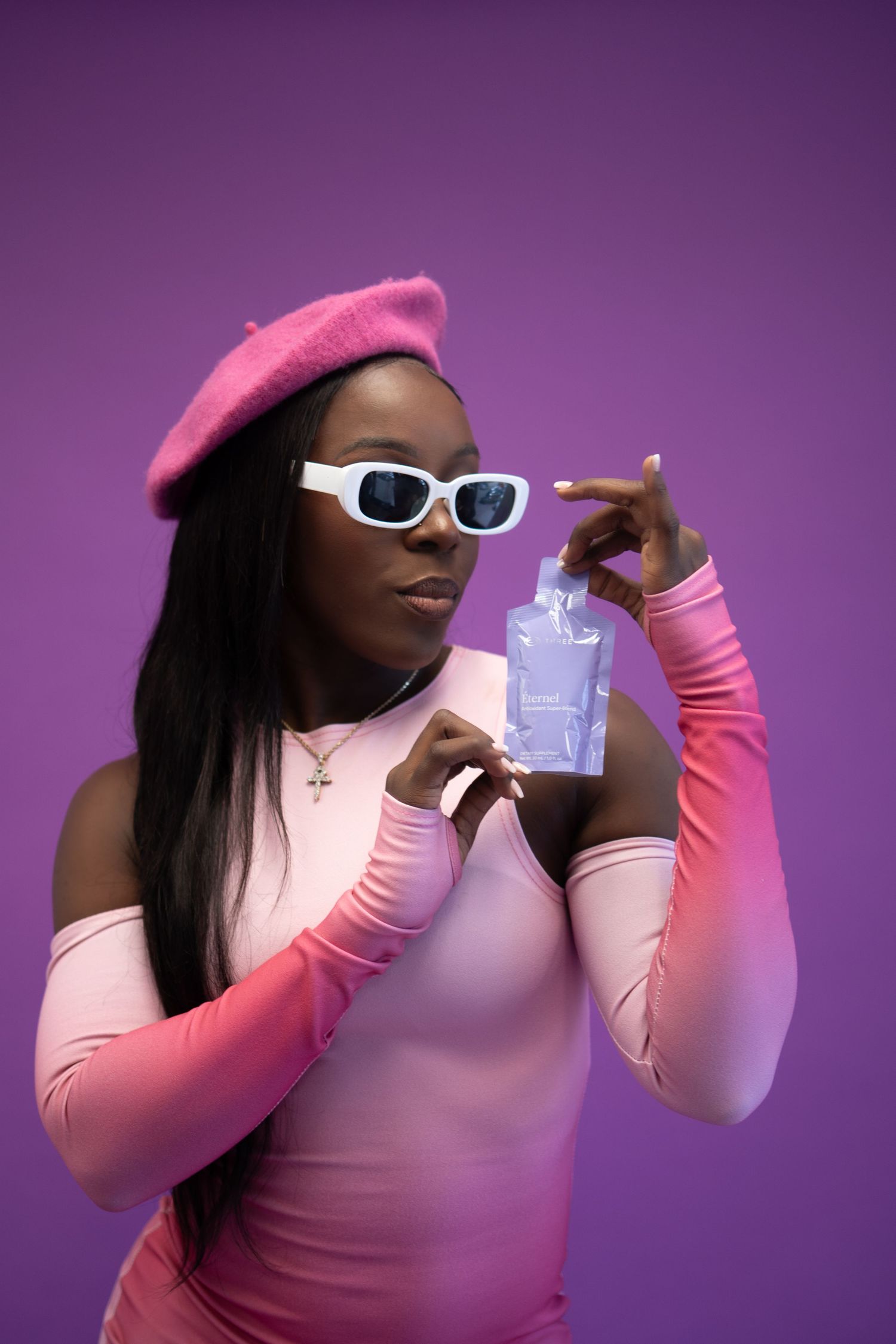 Stylish Black woman in pink beret and white sunglasses holding a lavender pouch against a purple background.