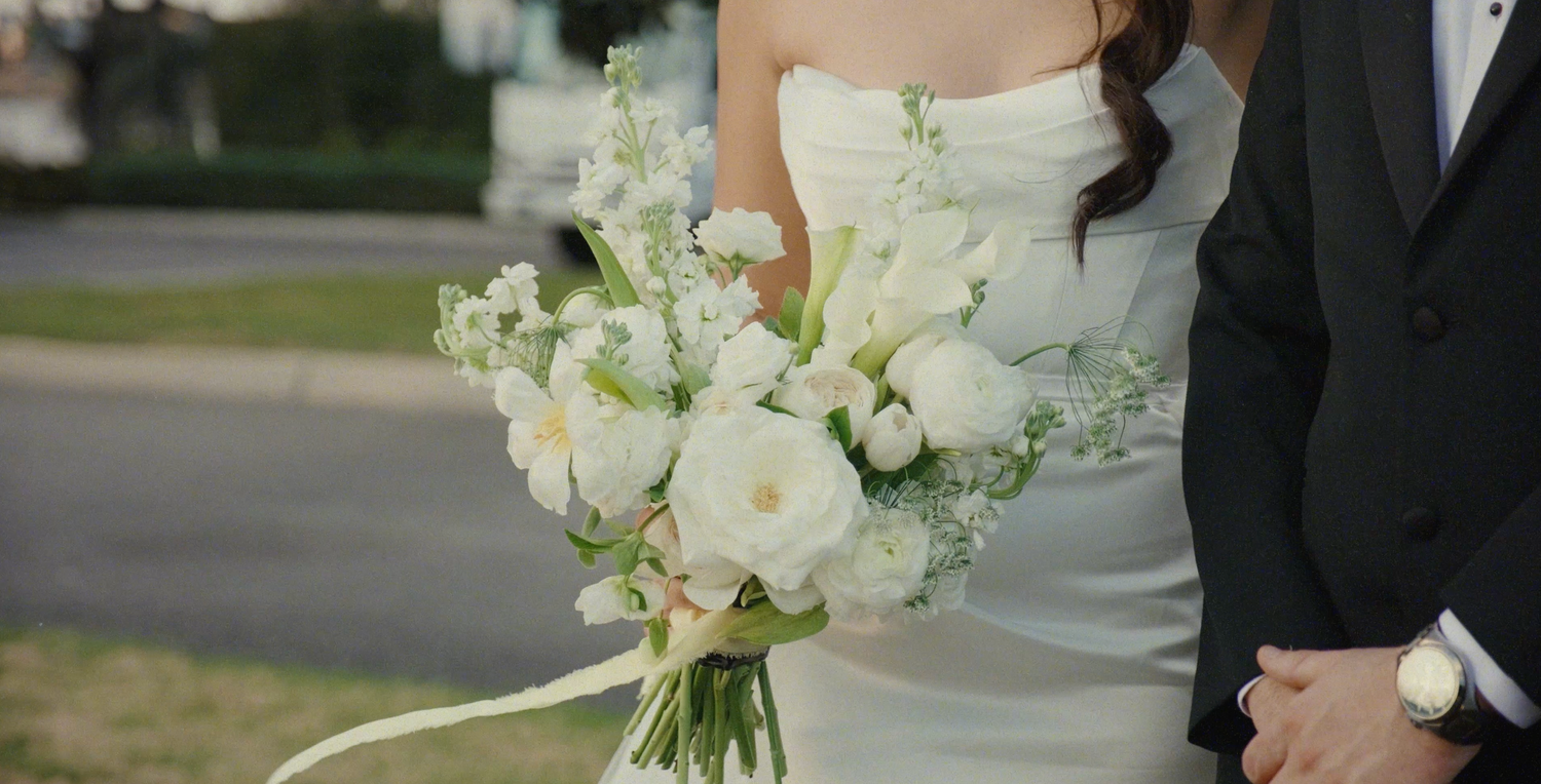 Bride in white gown holding elegant bouquet of white roses and greenery stands beside groom in black tuxedo.