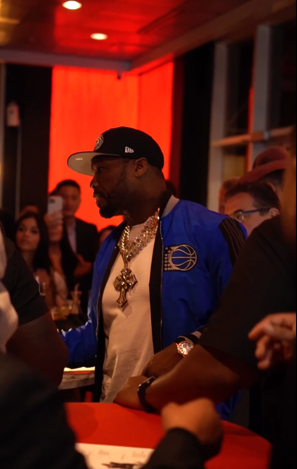 Man in blue varsity jacket, chain necklace, and cap stands at a crowded indoor event with red ambient lighting.