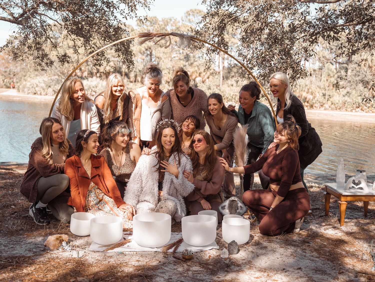 Group of women smiling around crystal singing bowls outdoors near a lake under trees.