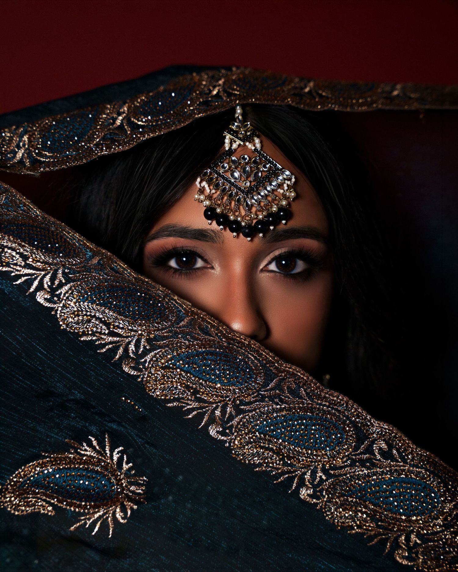 A woman in a teal embroidered dupatta with maang tikka jewelry, her eyes visible above the draped fabric.